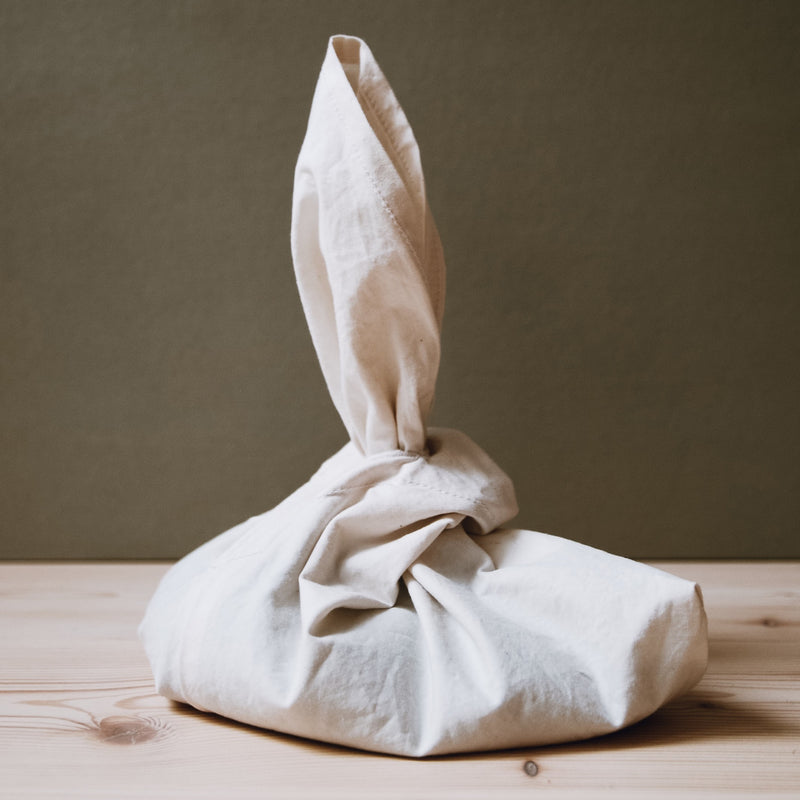 The Fill it by GoodGoods is a beige organic cotton fabric bundle resembling a reusable bag. Tied with an upward-pointing end, it rests elegantly on wood against an olive-green backdrop, ideal for non-packaging shopping.