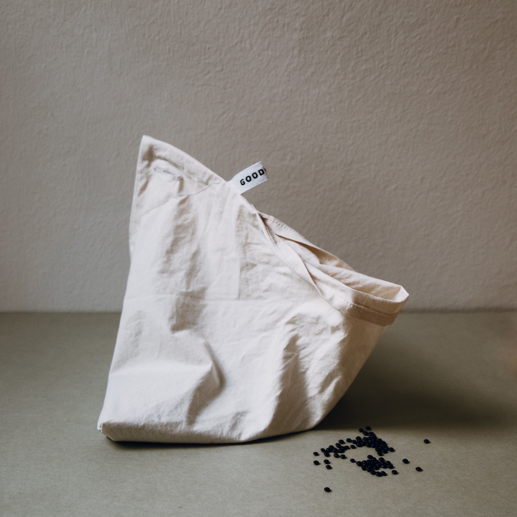 A Fill it bag by GoodGoods, crafted from white organic cotton, stands upright on a beige surface with some black lentils inside and scattered around. The tag promotes non-packaging shopping against a plain, light-colored wall.