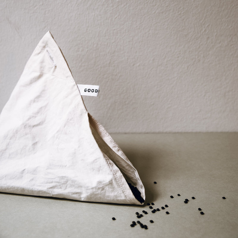 A triangular beige fabric bag made from organic cotton, branded GoodGoods and named Fill it, is adorned with small black beads spilling out of its opening. Set against a neutral background, this reusable bag combines sustainability and elegance, perfect for non-packaging shopping.