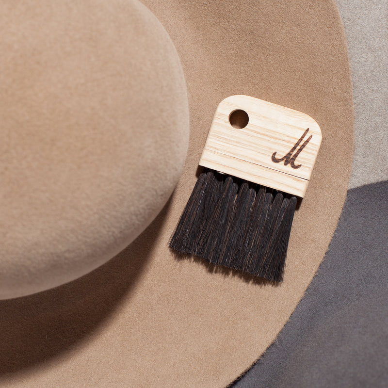 The Fuzz by GoodGoods is a beige felt hat featuring a small, portable brush with black horsehair bristles and an inscribed M on the handle, set against soft grey and beige surfaces.