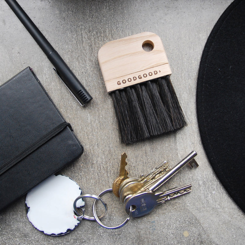 A closed black notebook, a pen, and a GoodGoods Fuzz ash wood horsehair brush occupy the desktop. Nearby, keys with a white round keychain rest on the gray surface, serving as a neutral backdrop for these casually arranged items.