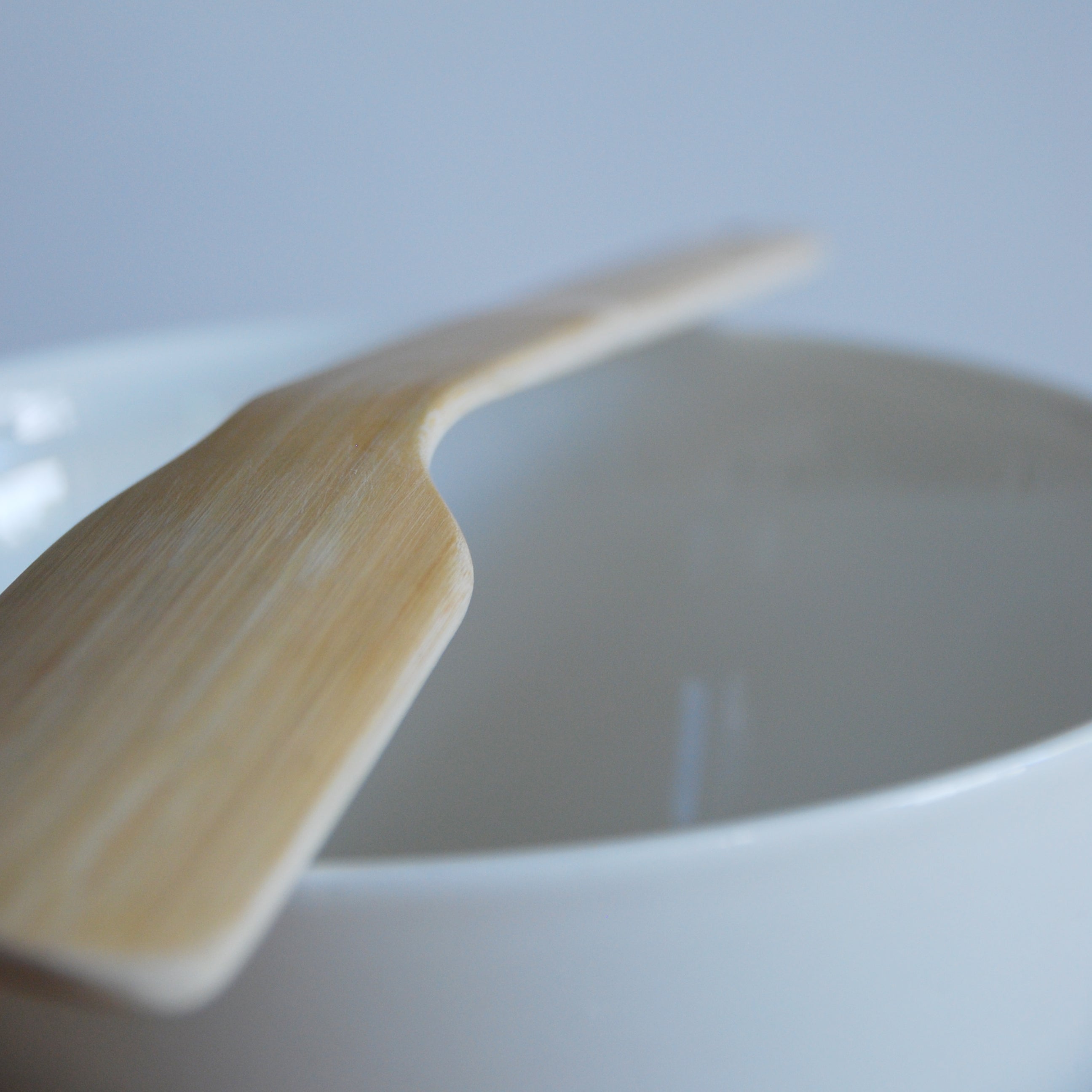 A GoodGoods Goodspoon, crafted from light bamboo, rests on the edge of a white bowl. The softly blurred background features subtle blue and gray tones.