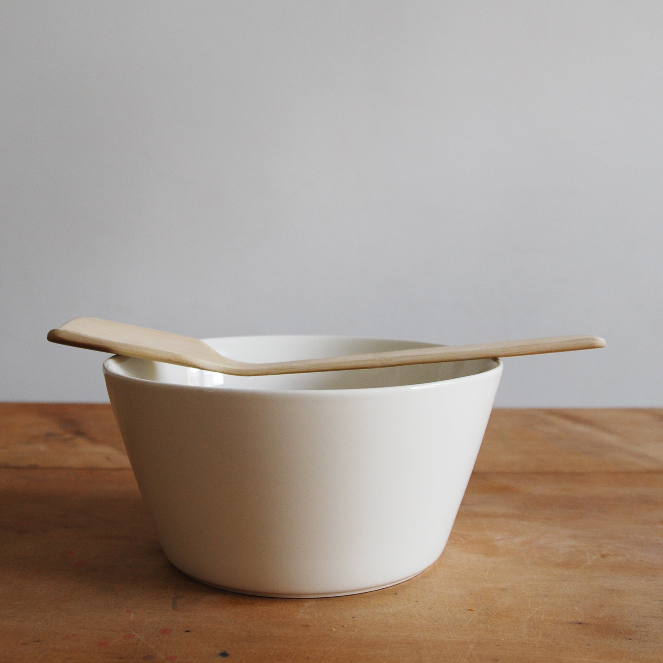 A Goodspoon by GoodGoods, a simple white ceramic bowl topped with a bamboo spoon, rests on a wooden surface. The softly lit background is plain.