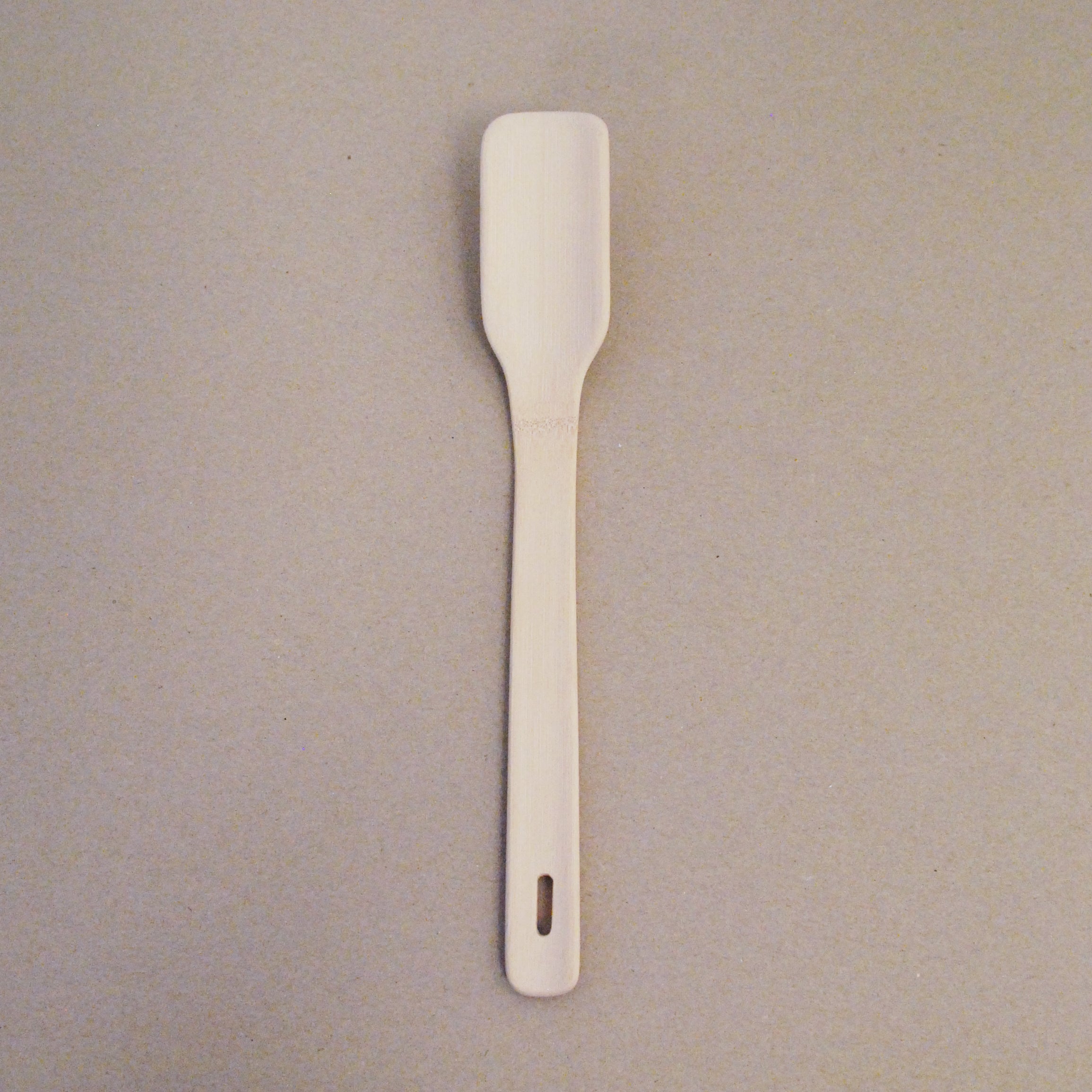 The GoodGoods Goodspoon is a bamboo-crafted wooden spatula with a rectangular head, long handle, and a small hanging hole, showcased on a light-colored surface.