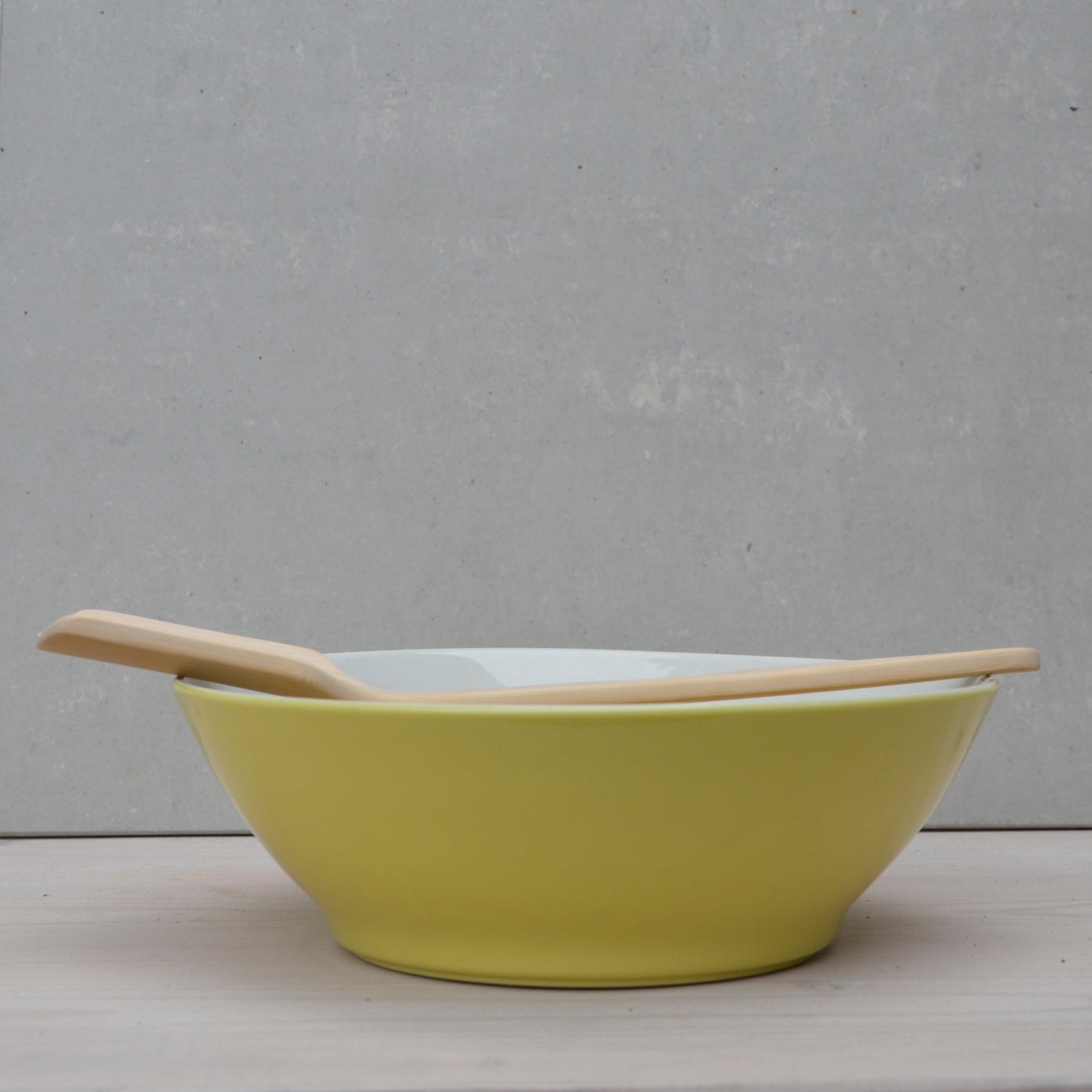 The GoodGoods Goodspoon bamboo spoon rests on the edge of a yellow bowl, placed on a light wooden surface with a plain gray background.