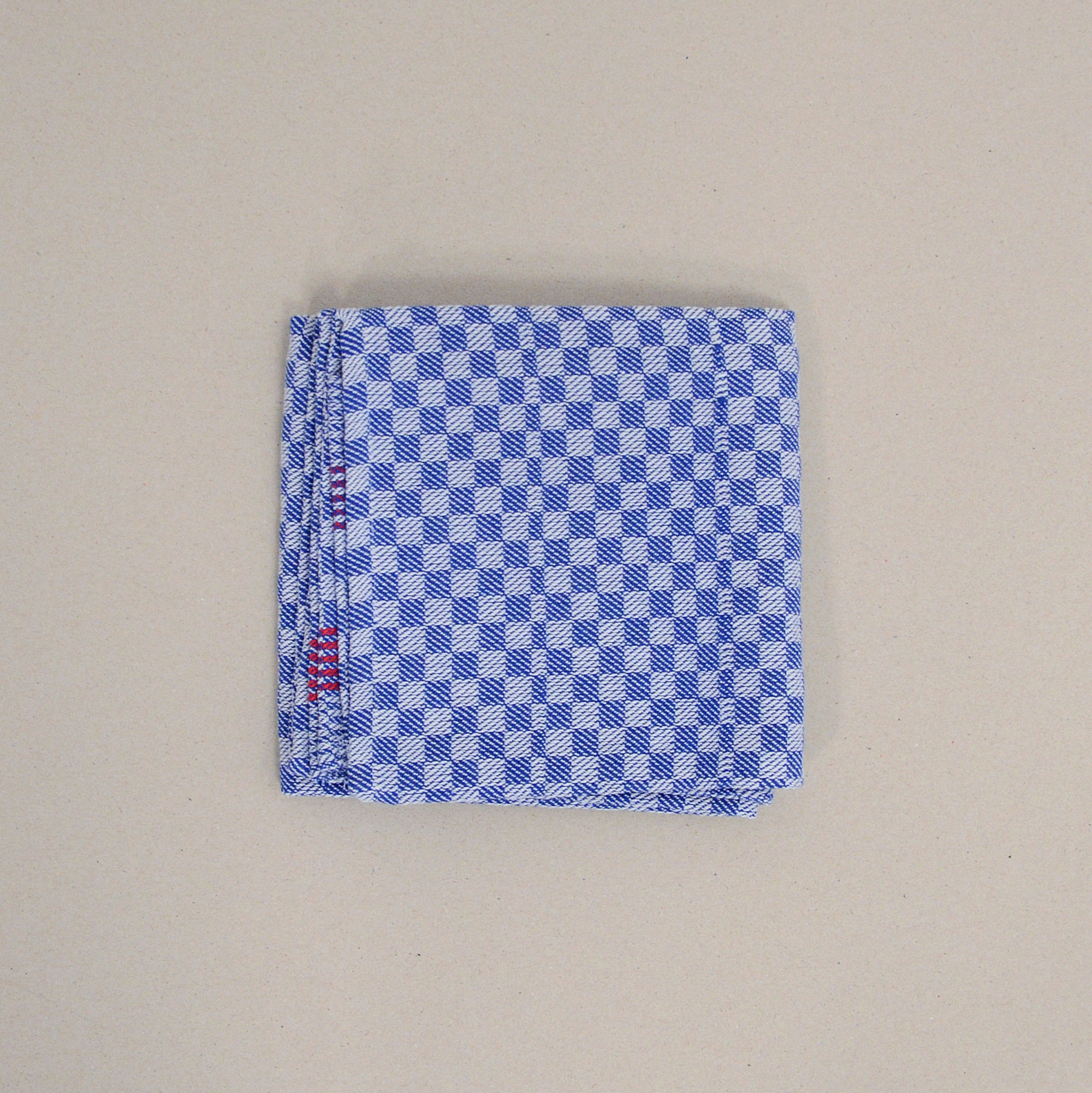 The product Dry by GoodGoods features a neatly folded cotton handkerchief in blue and white checkered pattern with a red detail at one corner, displayed on a plain beige background.