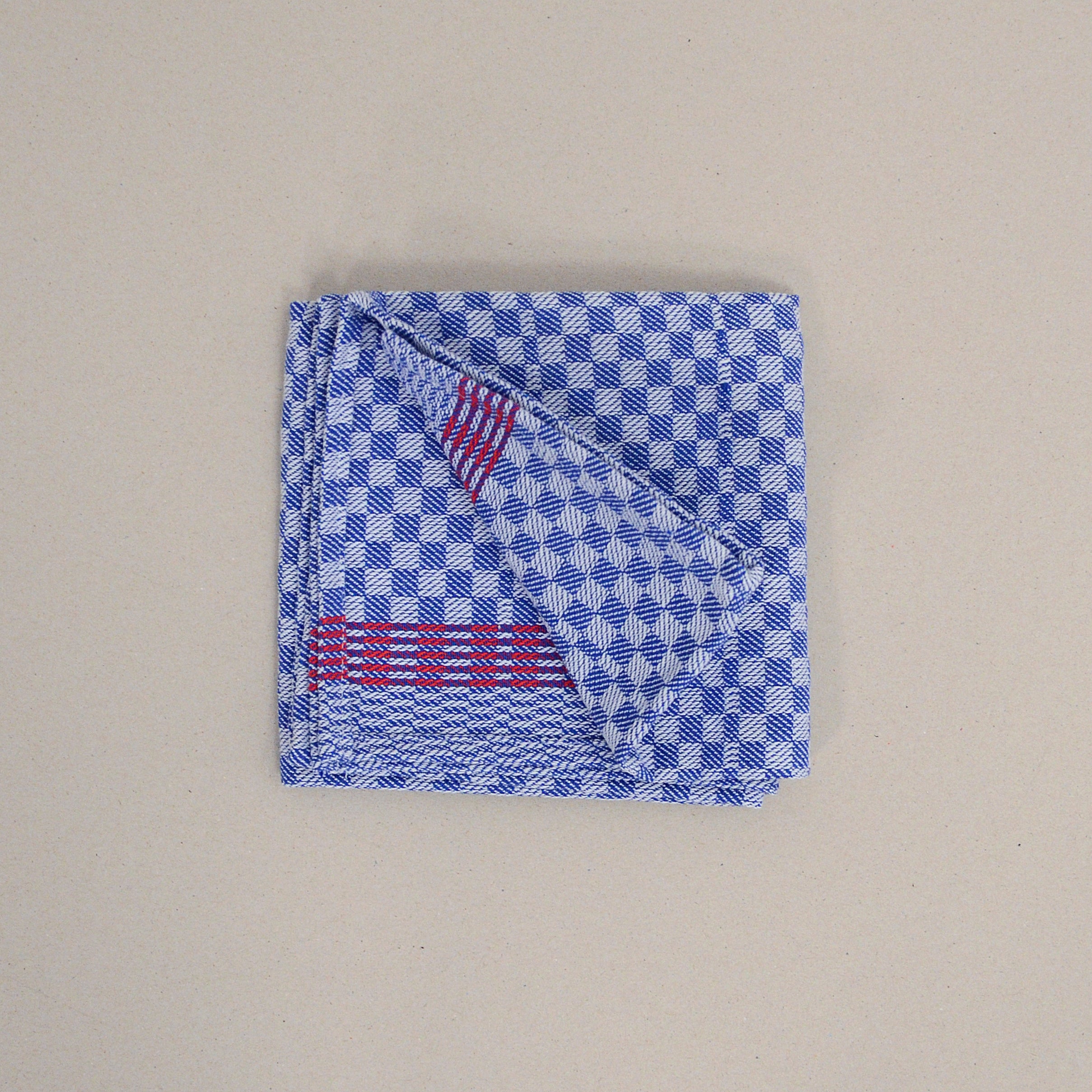 A neatly folded blue and white checkered GoodGoods cotton handkerchief, featuring red stripes on one corner against a beige background, exudes Drys quality.