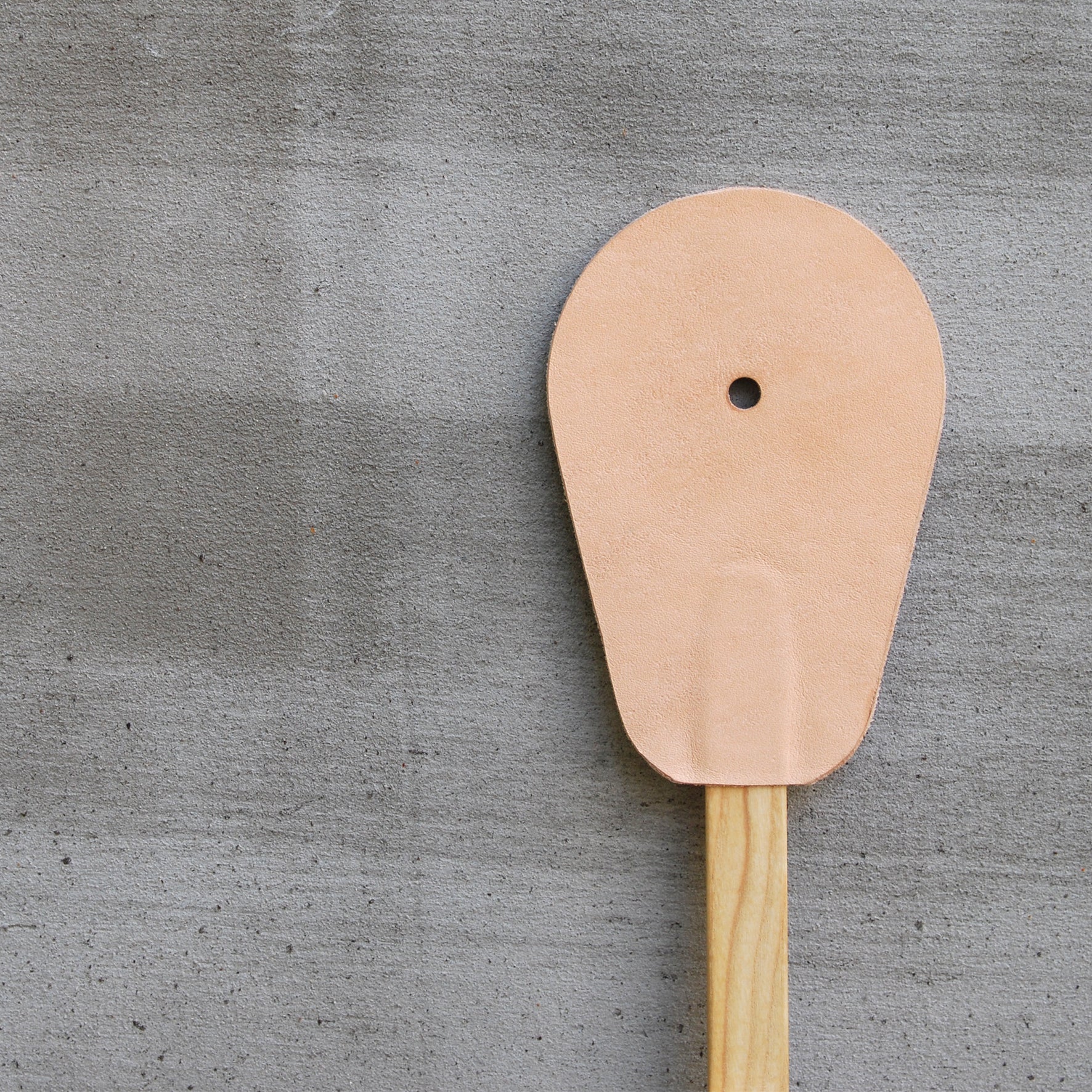 The GoodGoods Hit is a unique design featuring a round, flat leather paddle with a small central hole, attached to a wooden handle, reminiscent of a vintage fly swatter. It rests elegantly against the gray concrete surface.