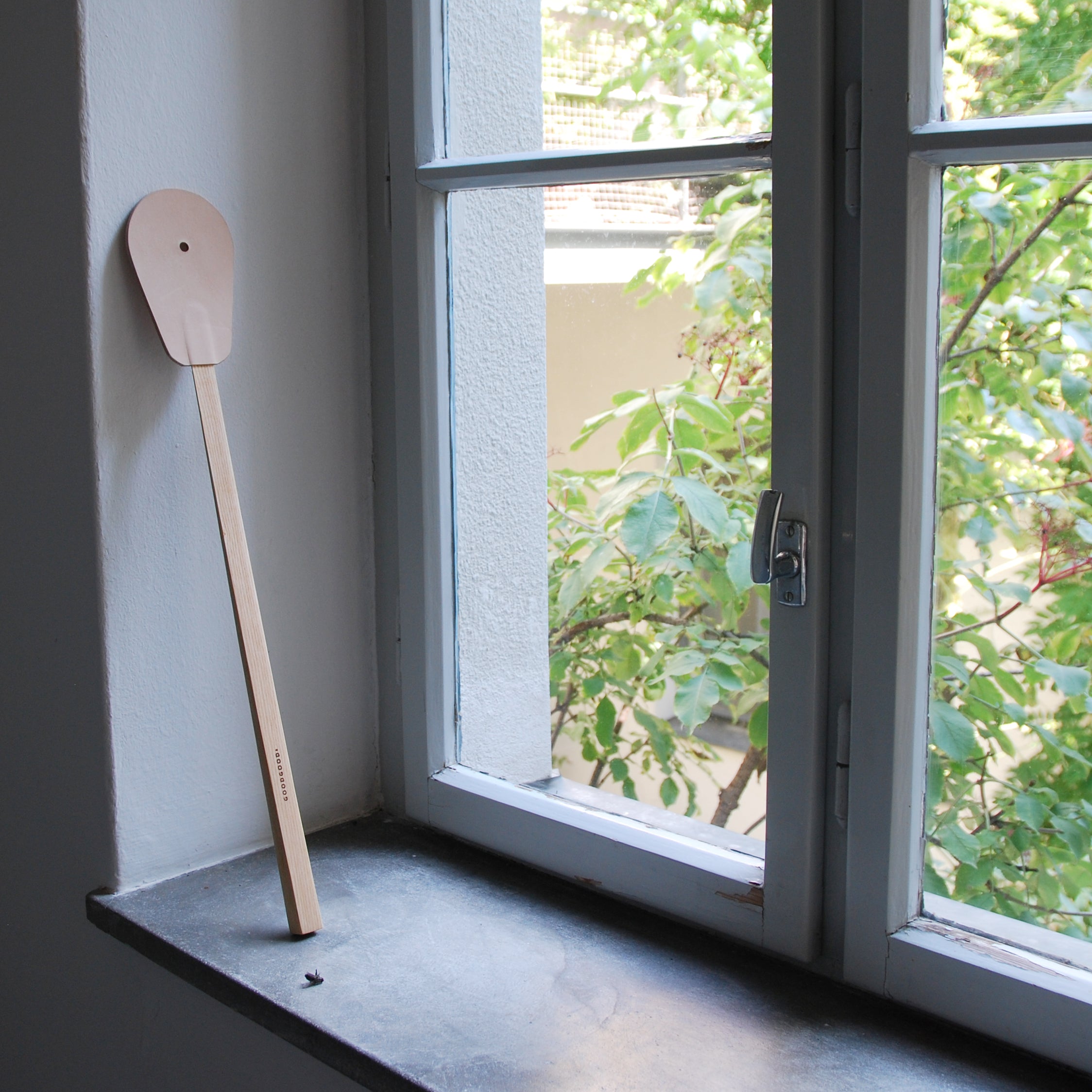 A GoodGoods Hit wooden spoon, resembling a fly swatter with its long handle, rests against the inside of a window frame next to a partially open window. Outside, green leaves and branches are visible, while the windowsill is made of dark stone.