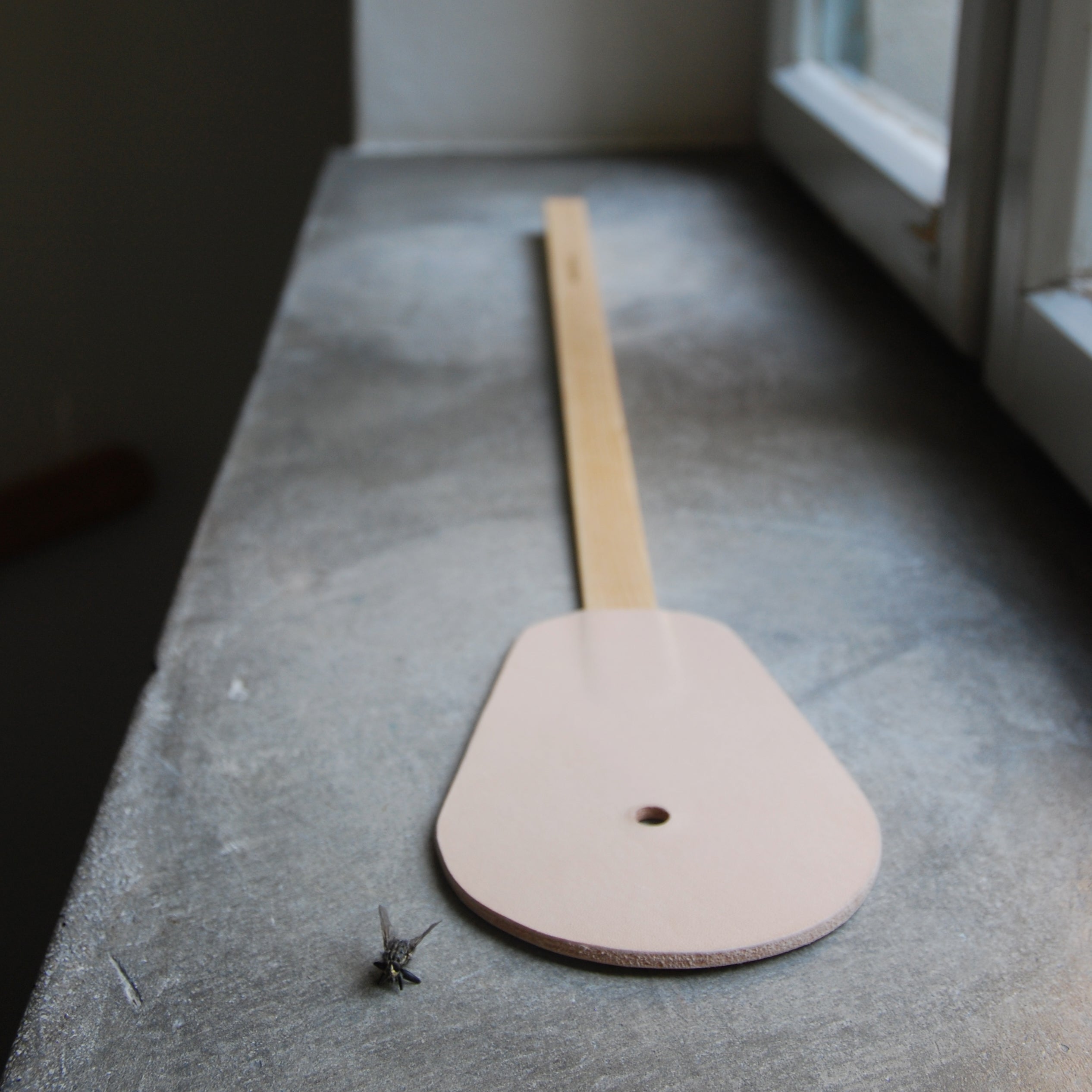 The GoodGoods Hit fly swatter, with its sophisticated wooden design and leather grip, rests elegantly on the windowsill. A single dead fly nearby attests to its precision, as soft light highlights its refined gray aerodynamics.