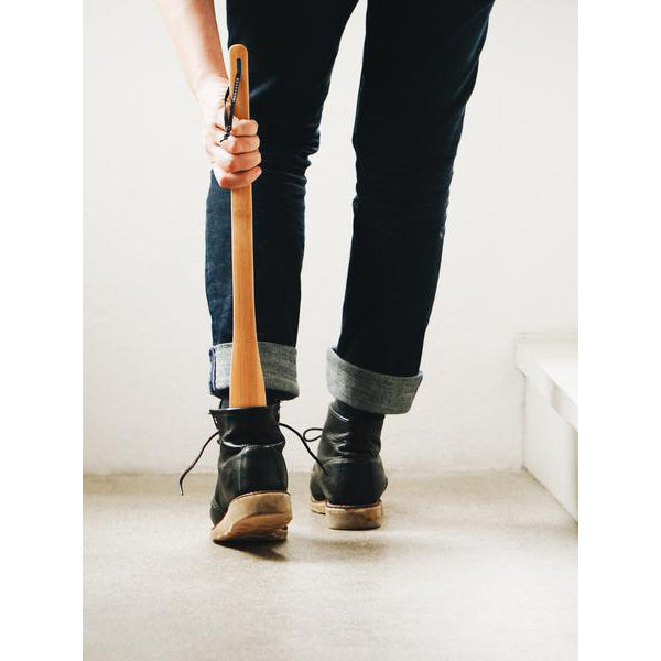 A person uses a GoodGoods On extra-long wooden shoehorn to slip into black boots. Clad in dark jeans, their legs and feet stand out against a light-colored floor.
