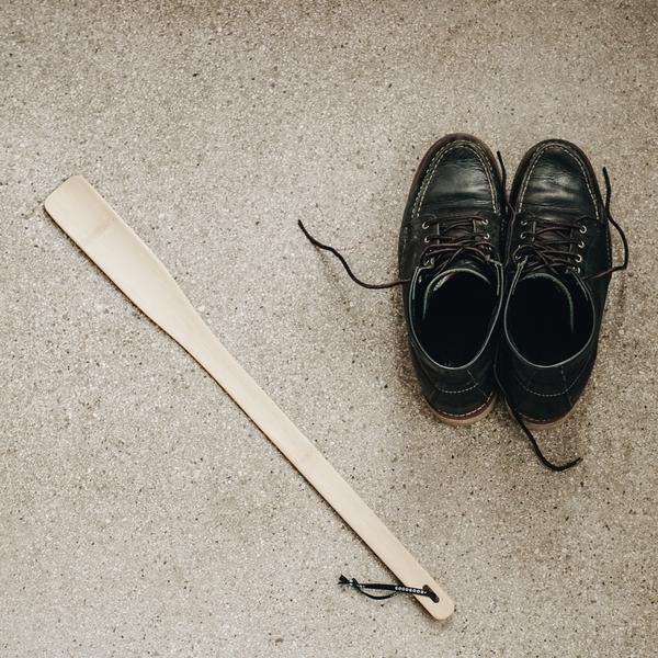 Black leather On shoes with untied laces rest on textured concrete, complemented by a GoodGoods extra long bamboo shoehorn with a small loop.