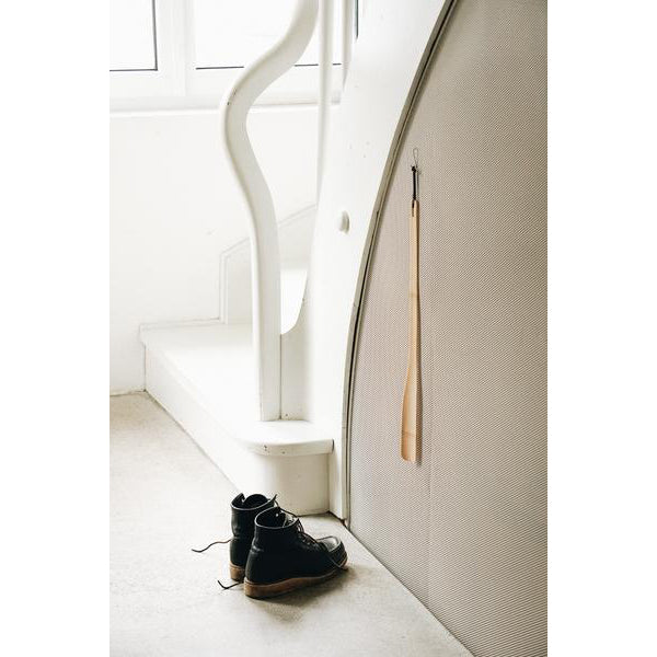A pair of black boots is on a light floor near a white staircase, with a sleek GoodGoods shoehorn from the On line hanging on the wall. The minimalist scene is illuminated by natural light streaming through the window.