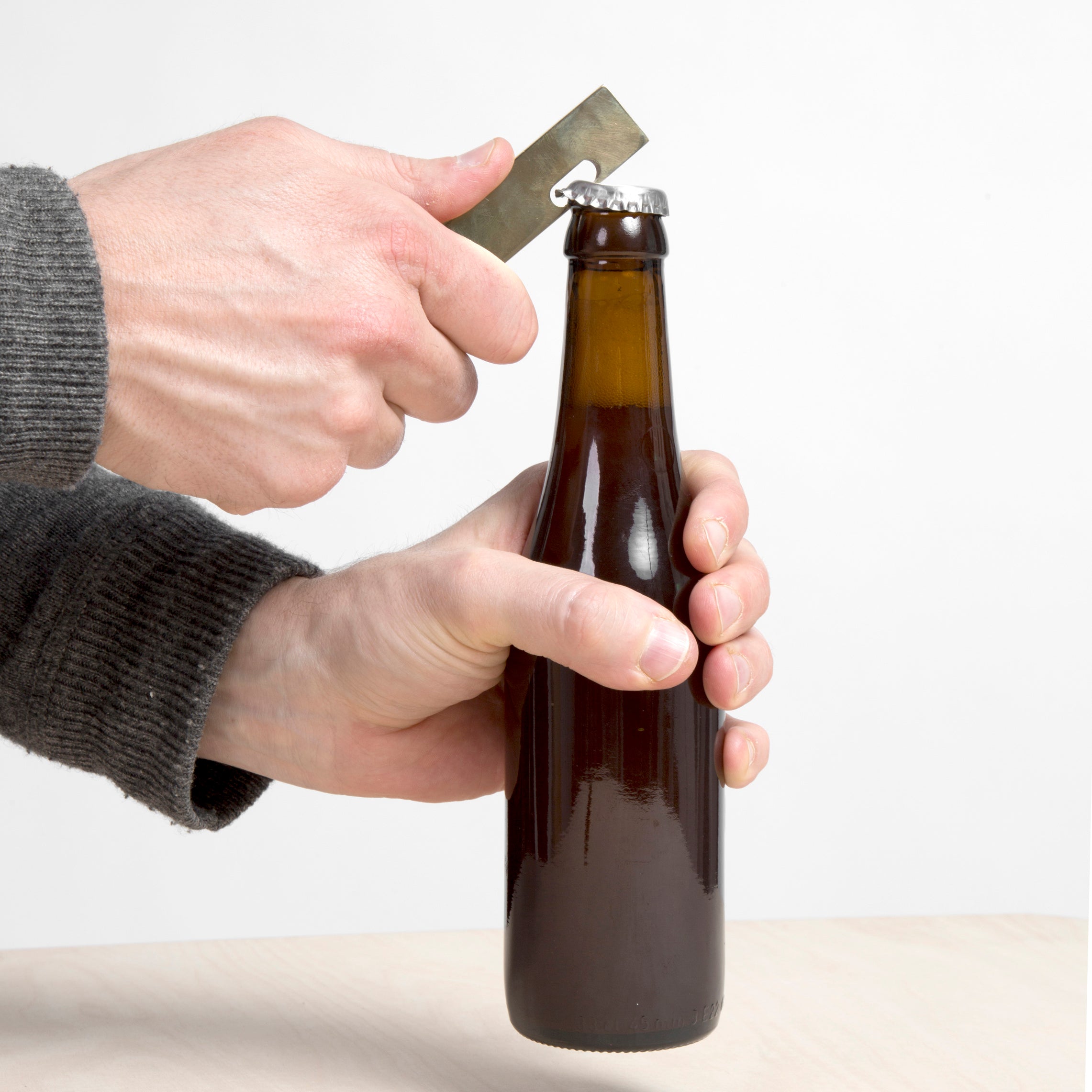 Hands using the GoodGoods Open brass bottle opener smoothly uncap a brown glass bottle. The person, dressed in a gray sweater, leans over the light surface, exuding sophistication and style similar to VANDASYEs design ethos.