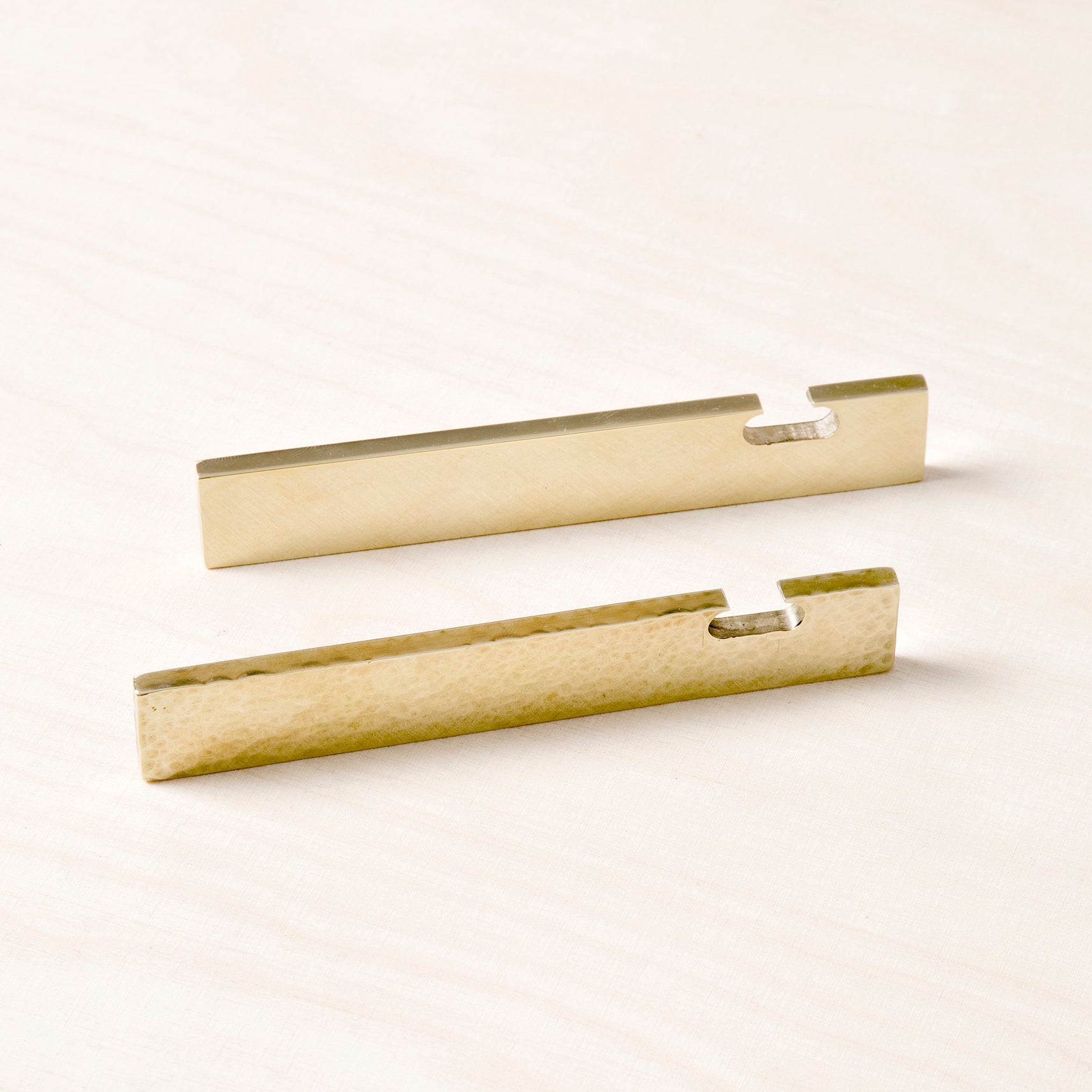 Two rectangular GoodGoods brass Open bottle openers rest on a light wooden surface; one has a smooth finish while the other features a hammered texture. Each opener includes a small notch for easy bottle cap opening.