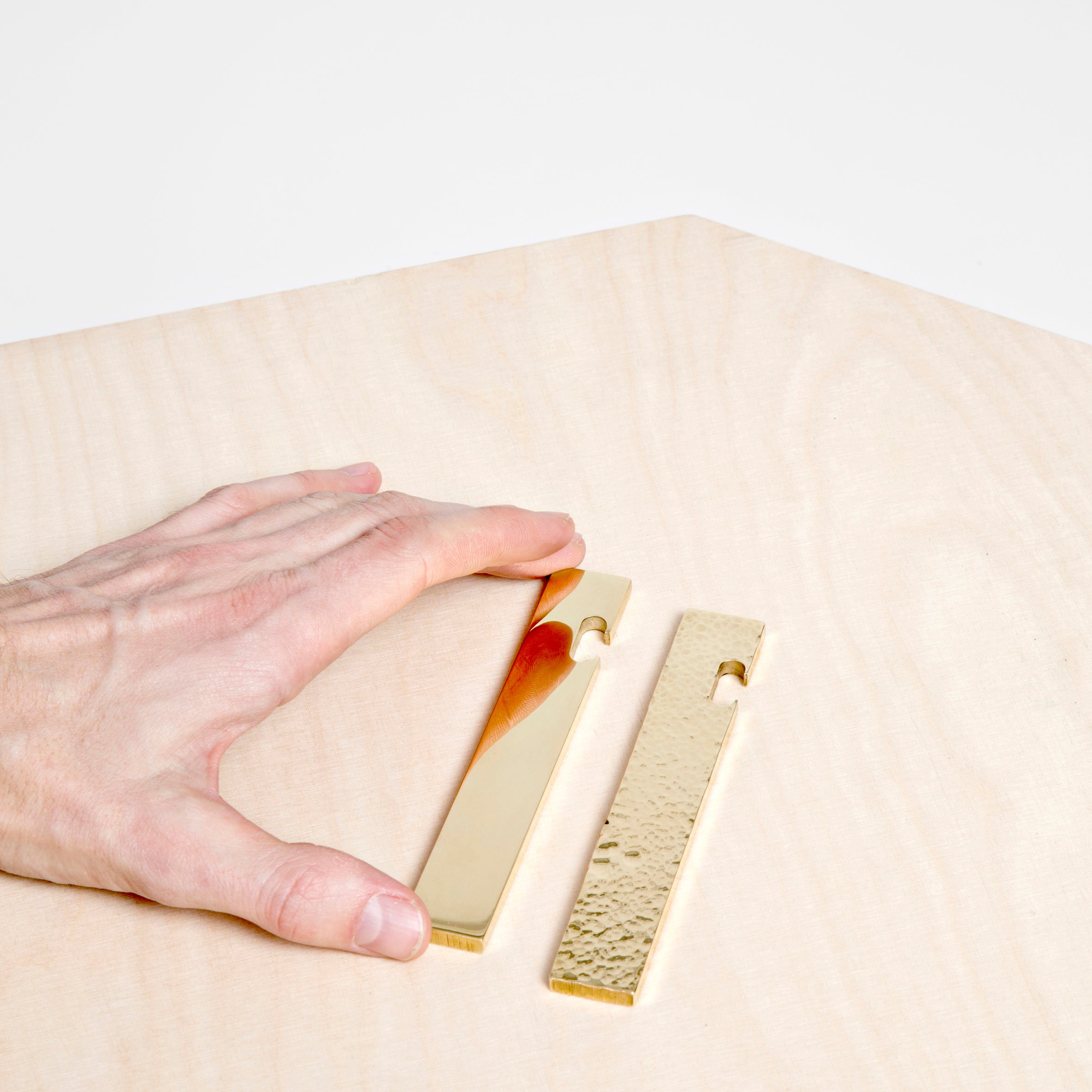A hand holds one of two rectangular, brass-colored Open bottle openers by GoodGoods on a light wooden surface. One is smooth while the other has a textured finish. Each opener features notches at one end, combining elegance and functionality.