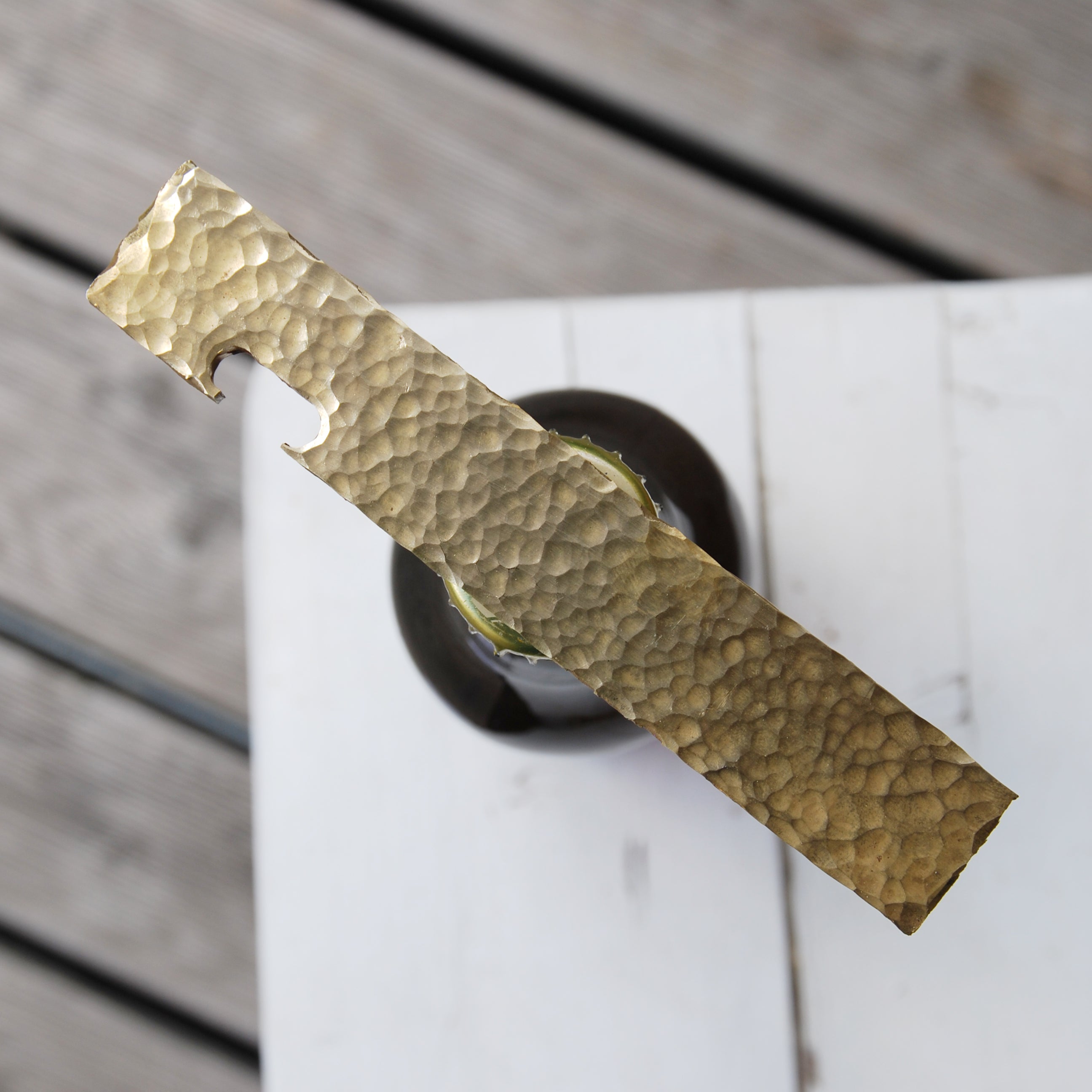 A close-up of a GoodGoods Open bottle opener with a hammered brass finish, positioned atop a partially opened dark glass bottle on a wooden surface. The opener stands vertically with the cap peeking out beneath it.