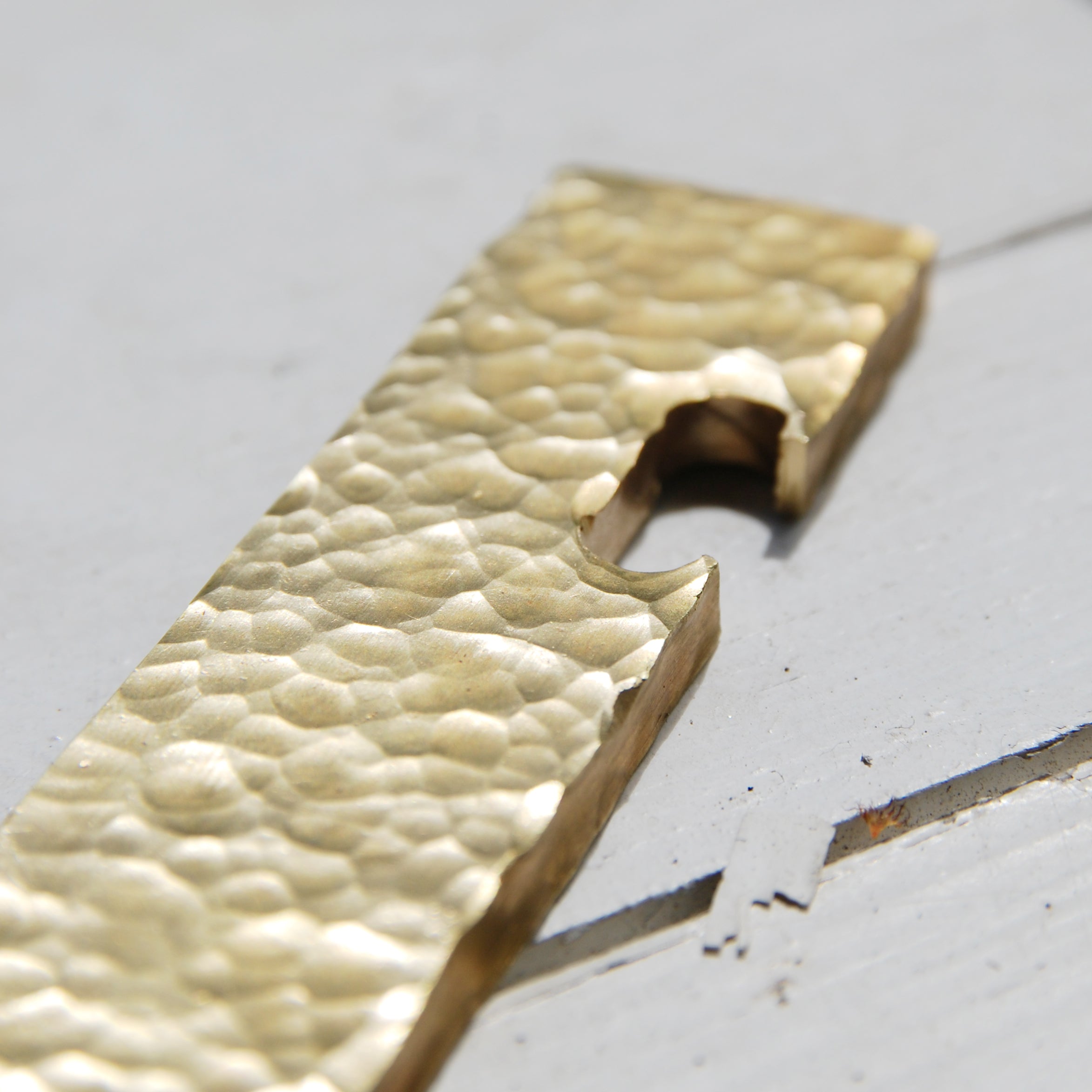Close-up of a brass rectangular object called Open from GoodGoods, featuring a notched cutout like a unique bottle opener, resting on a light gray surface with visible seams.