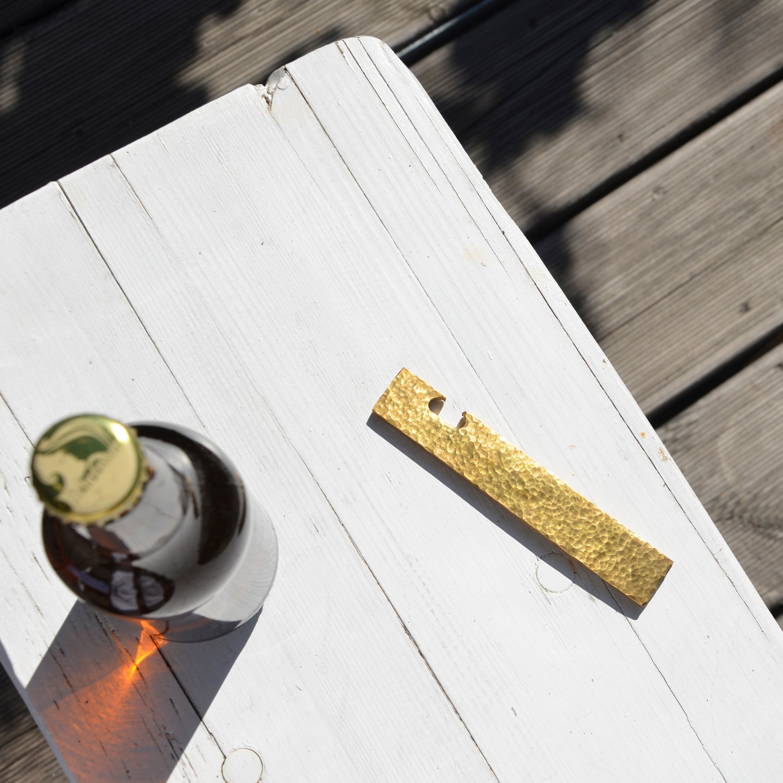 A brown bottle with a shiny gold cap rests on a white wooden surface next to a textured brass GoodGoods Open bottle opener, creating shadows that enhance the minimalistic and rustic setting.