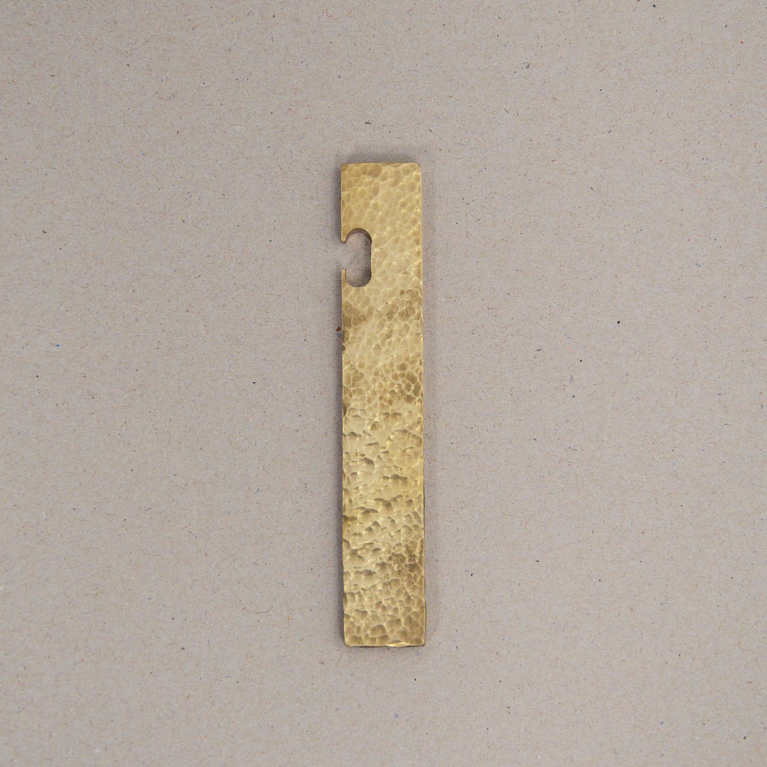 A rectangular, textured brass strip subtly branded with GoodGoods features a curved notch on the top edge. Elegantly placed vertically against a plain beige background, it hints at its dual function as a stylish bottle opener and is named Open.