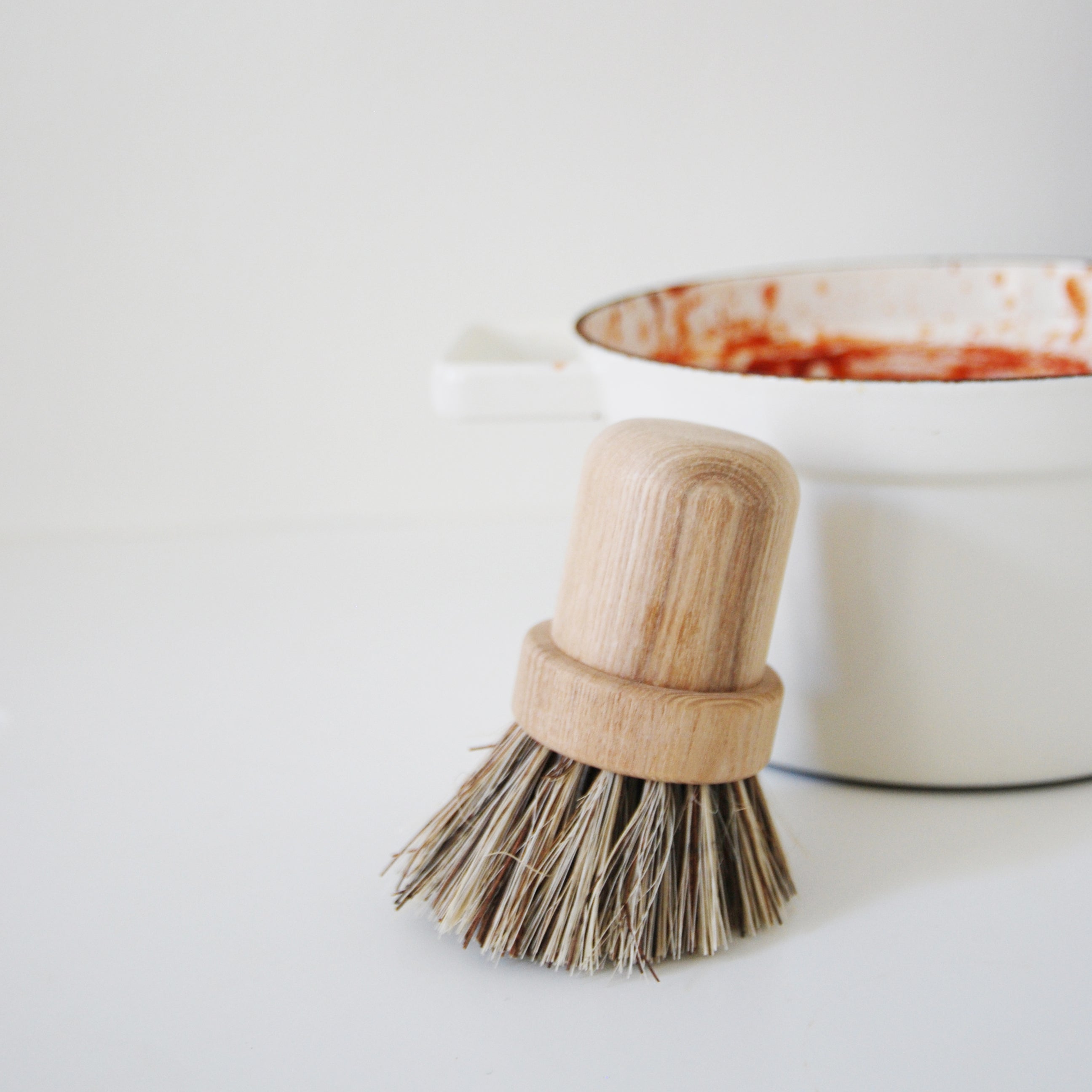 The Pisa pot brush by GoodGoods, made from ash wood with palm fiber bristles, stands on a white surface next to a white enamel bowl of red sauce, set against a neutral background.