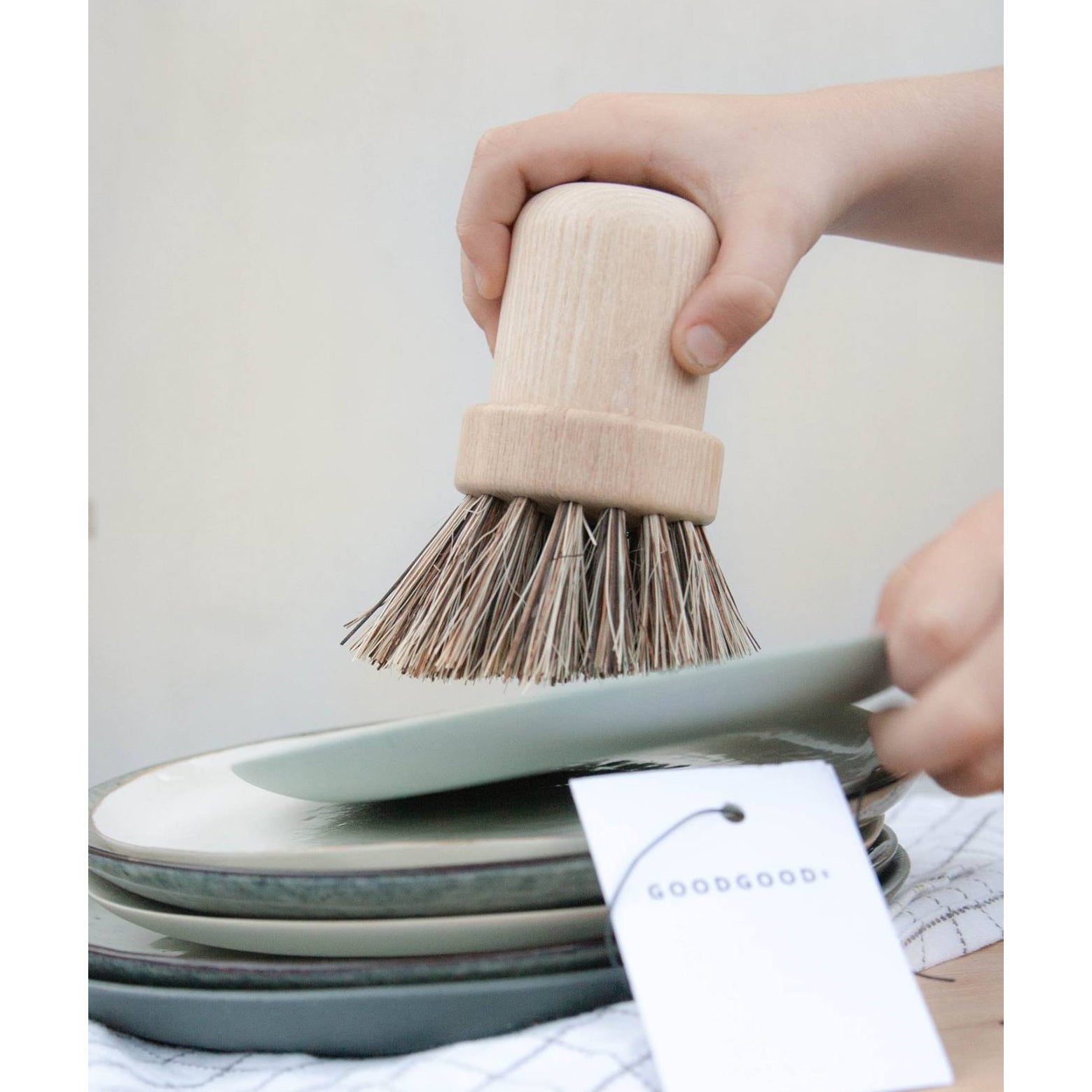 A hand uses the Pisa brush from GoodGoods, featuring an ash wood handle and natural palm fiber bristles, to scrub a stack of ceramic plates. A label hangs from one plate, set against a neutral background with soft lighting.