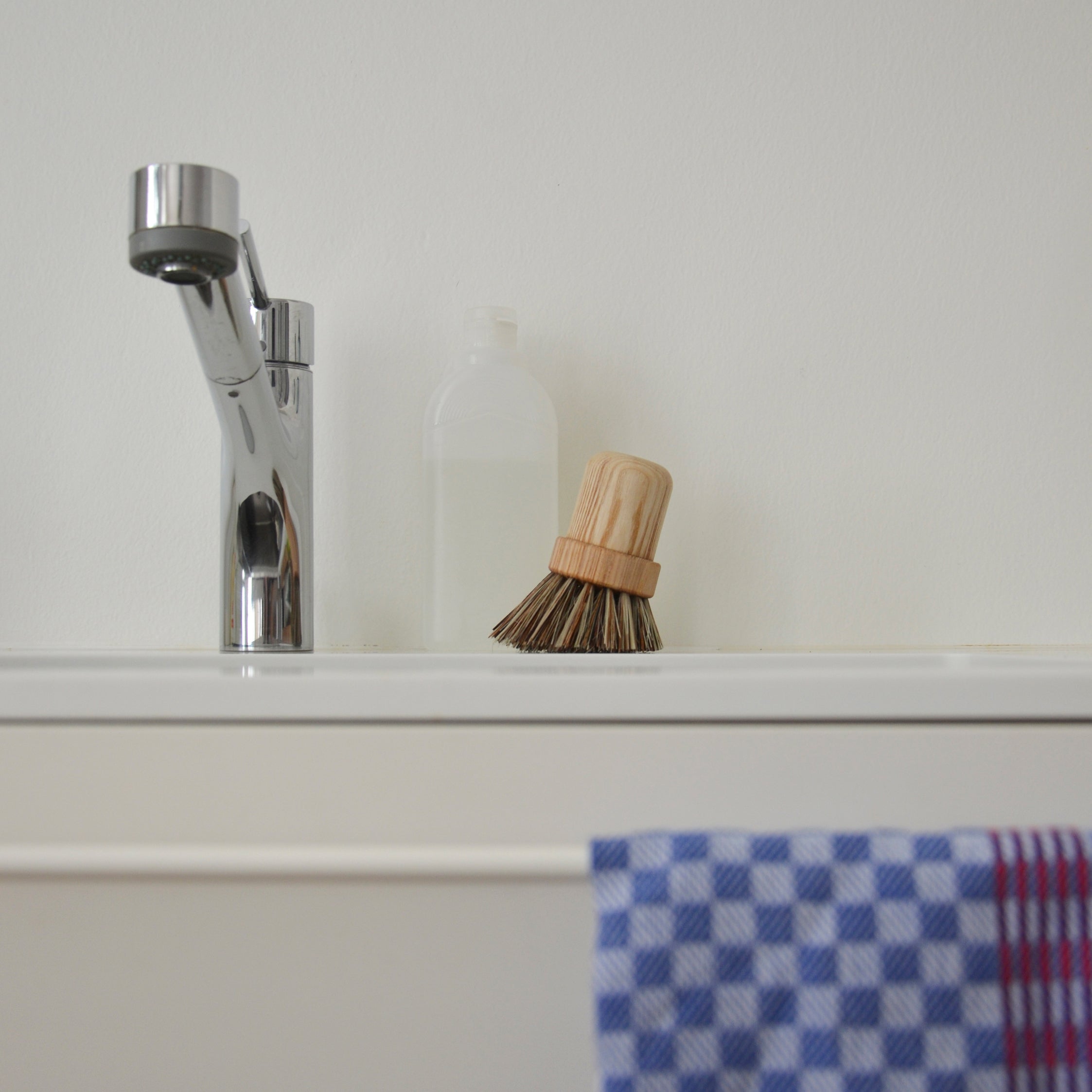 The Pisa white sink by GoodGoods, featuring a sleek modern chrome faucet, is paired with an ash wood pot brush on the edge. A small clear bottle accompanies a blue and red checkered cloth adorning the scene against a simple white wall, highlighting its understated elegance.