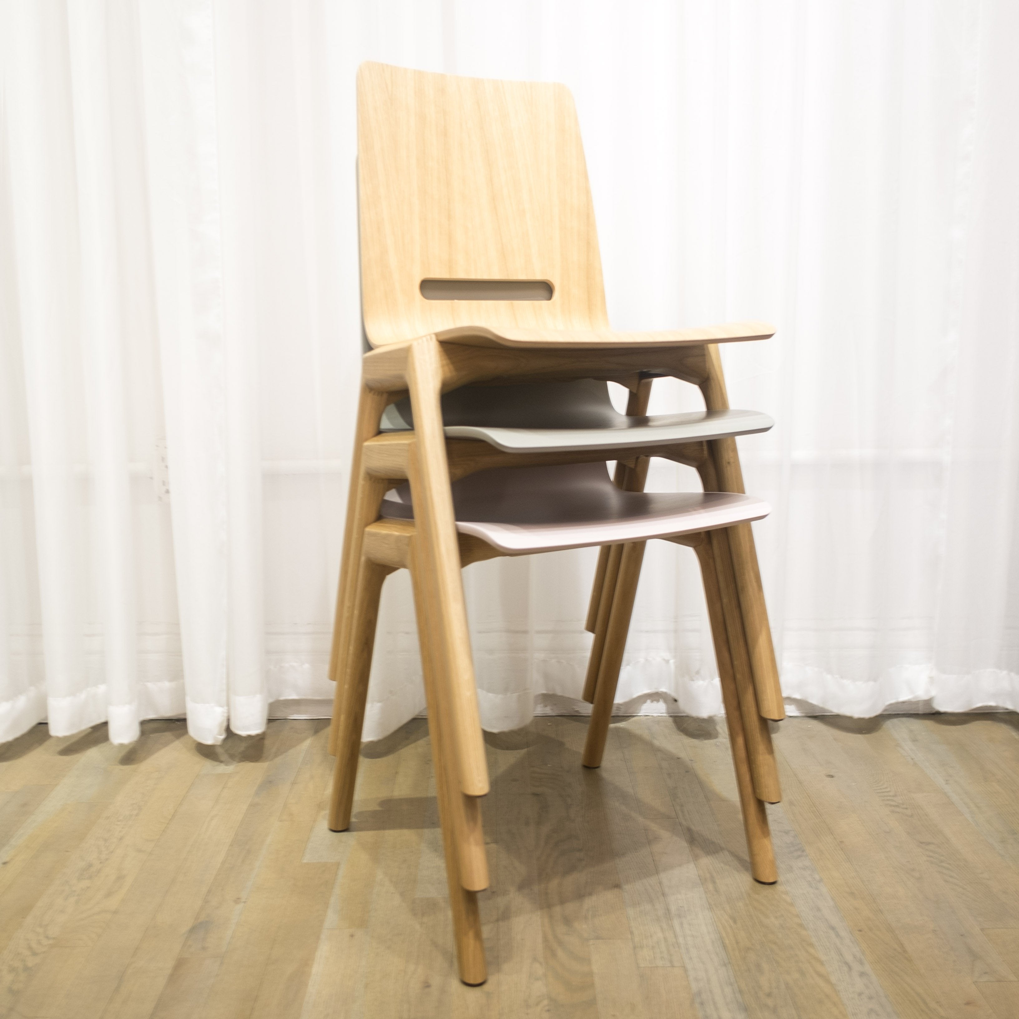 The Forum 2S chairs by Hussl feature A-shaped legs and light wood frames. The top chair has a darker tone, while those beneath have lighter molded ply seats and backrests. Stacked against a white curtain on a wooden floor, these stackable timber chairs exude contemporary elegance.