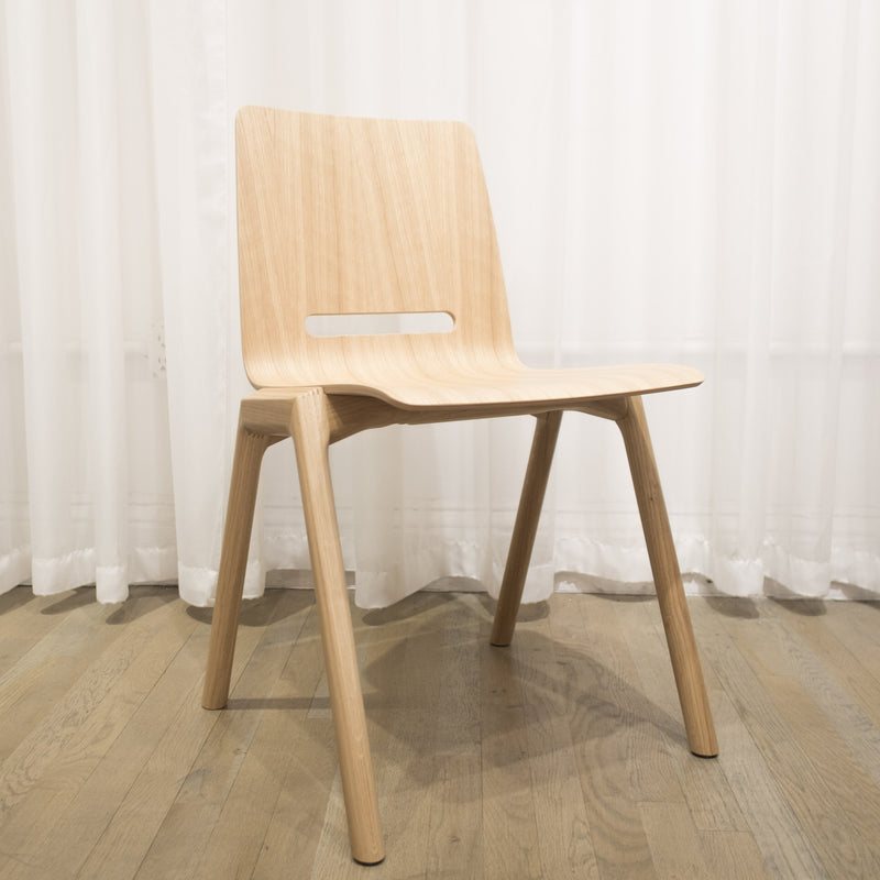 The Hussl Forum 2S - Sale, a modern and minimalist wooden chair with A-shaped legs and a smooth, light finish, rests on hardwood. It features a curved seat and backrest with a narrow horizontal cutout against sheer white curtains.