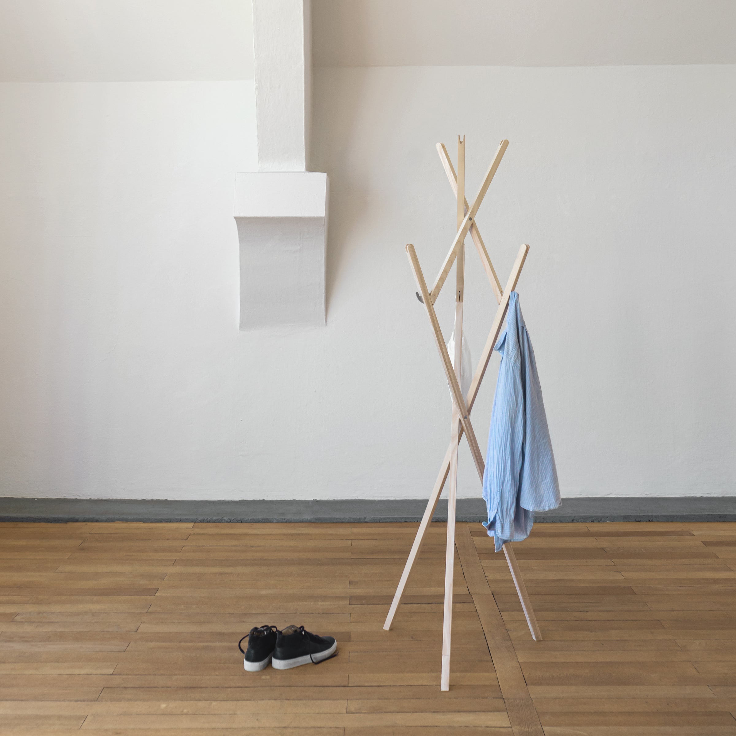 A minimalist room showcases the Moormann Hut ab adjustable coat rack displaying a blue shirt, while black shoes lie on the wooden floor. The plain white wall and angled ceiling enhance the open and airy ambiance.