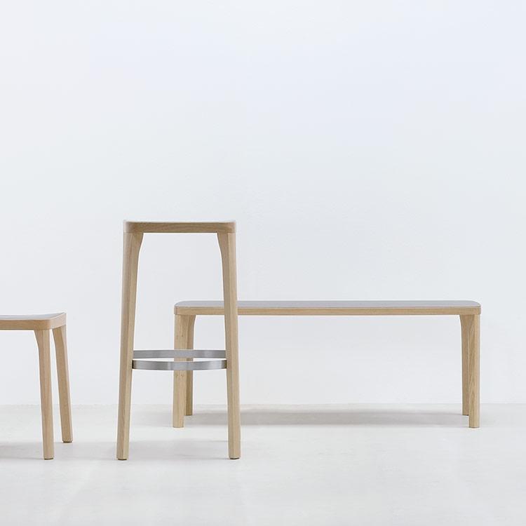 The minimalist furniture arrangement features a wooden stool, a sleek high stool with metal support, and an ST6 Bench from Hussl. Set against a plain white background, it evokes an Austrian coffee house vibe with its modern aesthetic and focus on natural wood tones.