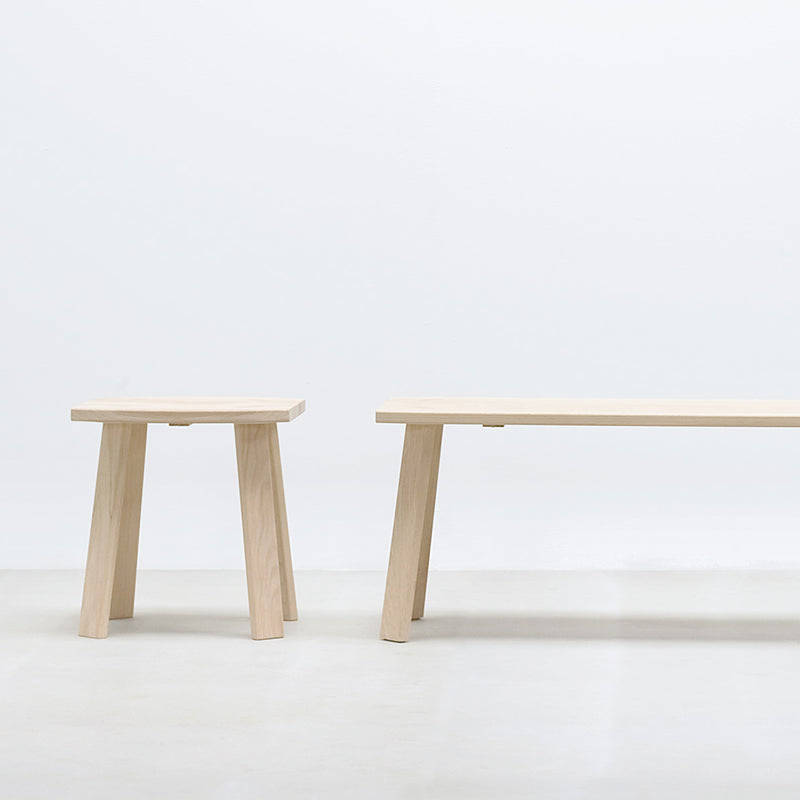 The Alpin Bench by Hussl, with its minimalist design by EOOS, features a small square stool and a rectangular bench with angled legs. Set against a white backdrop, it captures the elegance of Alpine furniture.