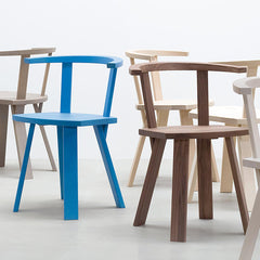 The Alpin Armchair collection by Hussl, inspired by minimalist design, features distinctive curved backrests and armrests. Available in blue, brown, and light wood, they are elegantly arranged on a smooth white floor in a bright room.