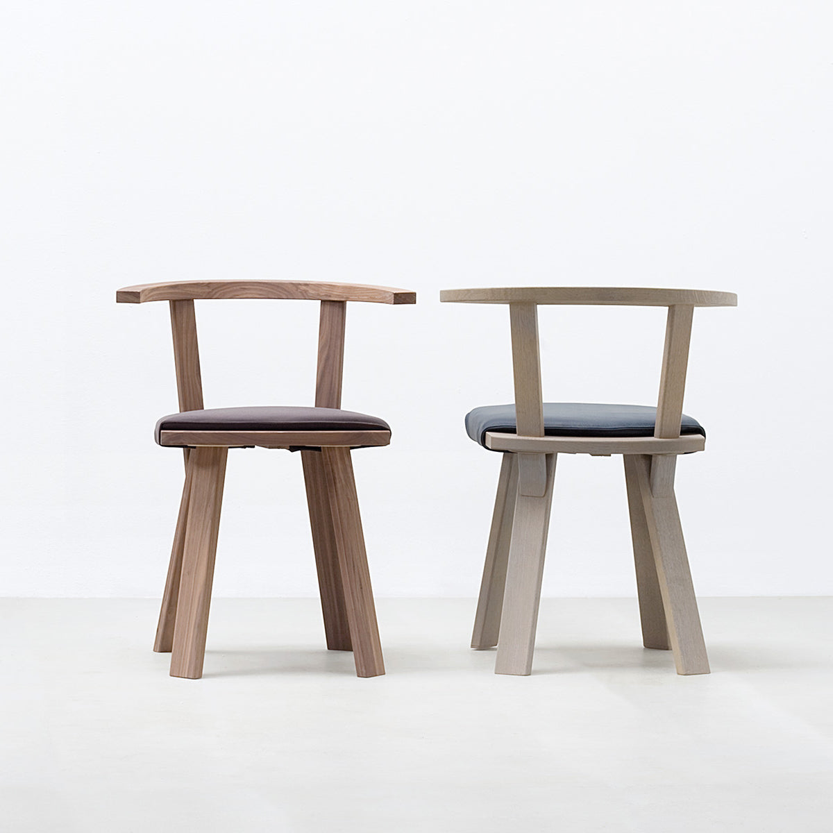 Two Hussl Alpin Armchairs feature wooden frames and upholstered seats: one in natural wood with a dark brown cushion, the other in light wood with a gray cushion. Both chairs elegantly contrast against a plain white background on a light gray floor.