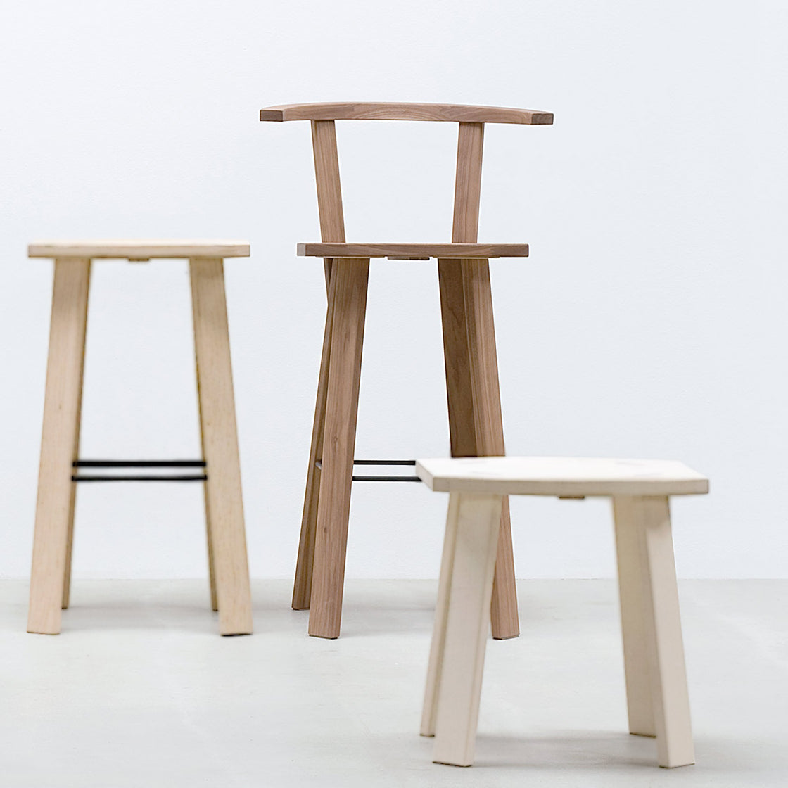 Three wooden Hussl Alpin stools of varying heights sit on a light gray floor against a white wall. The tallest one has a backrest, while the others offer flat seats. Each stool features a minimalist design.