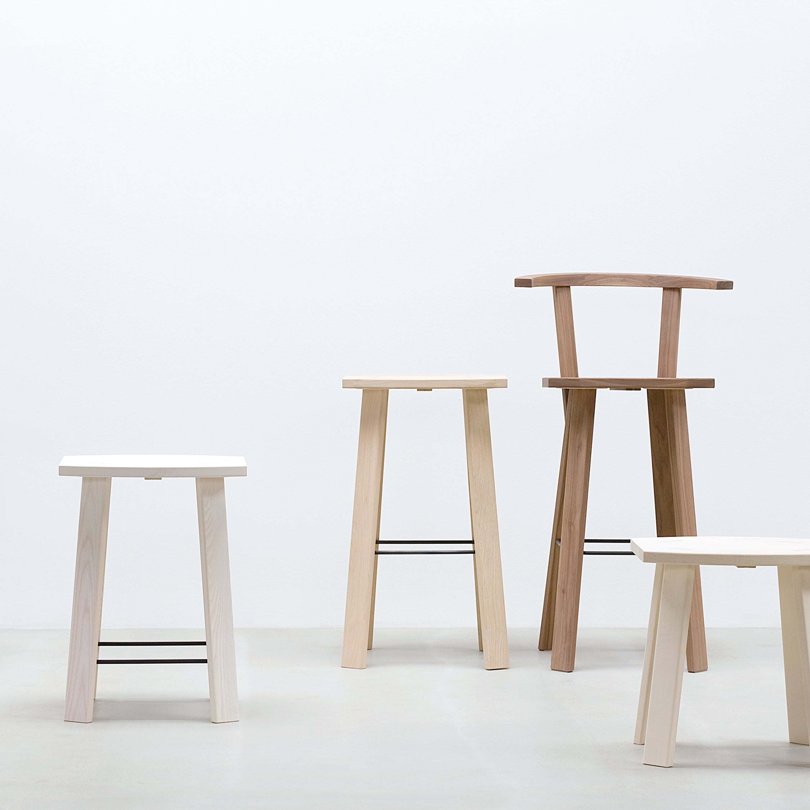 A minimalist setting features four Hussl Alpin Barstools without backrests on a light grey floor against a plain white wall. These stools, inspired by natural wood finishes, display simple lines and black metal footrests in varying heights and styles.