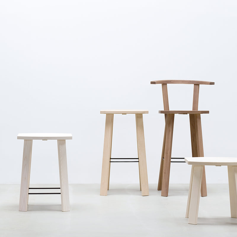 A minimalist setting features four Hussl Alpin Barstools without backrests on a light grey floor against a plain white wall. These stools, inspired by natural wood finishes, display simple lines and black metal footrests in varying heights and styles.