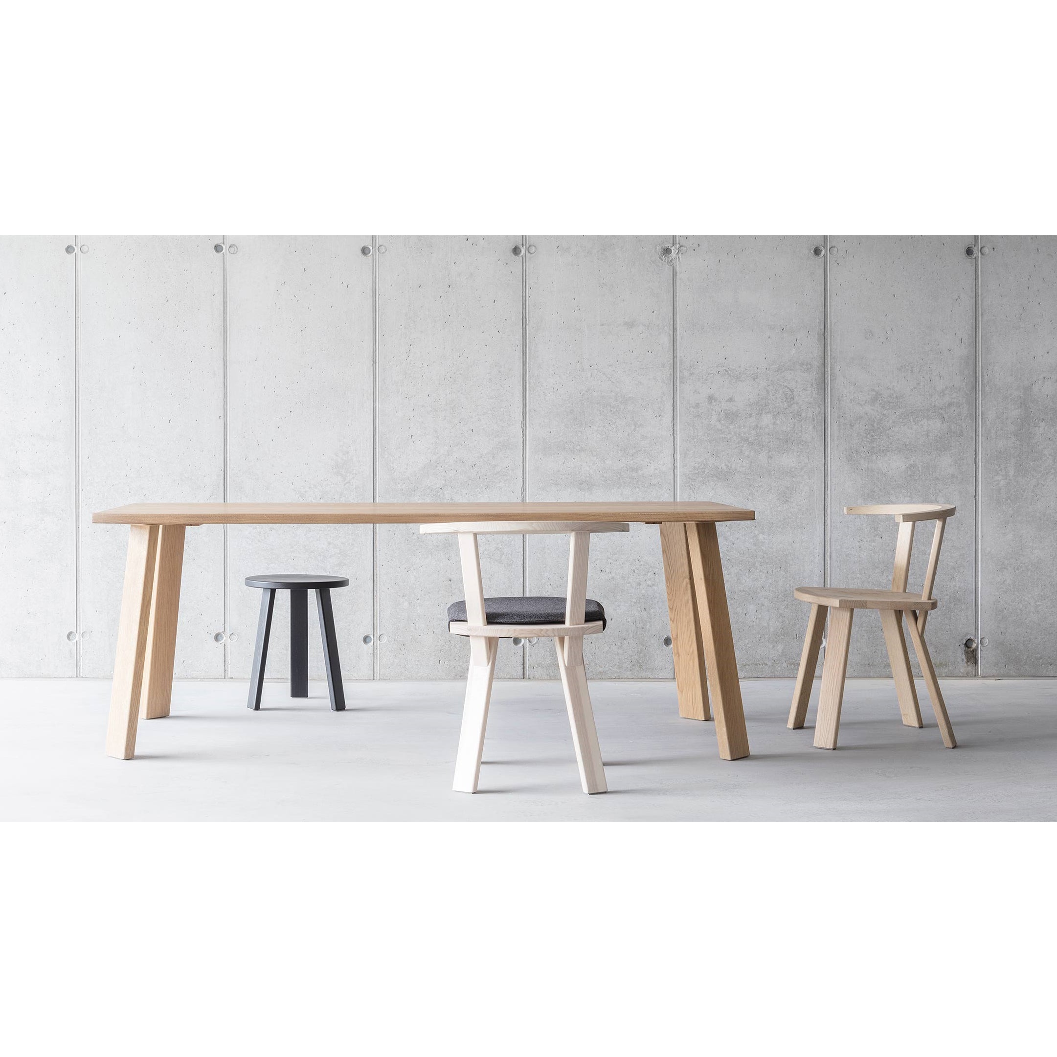 The Alpin Table, by Hussl, paired with two contrasting chairs—one light wood and one dark-cushioned—and a small black stool, creates an elegant EOOS look against a minimalist concrete backdrop, epitomizing the Alpine aesthetic.