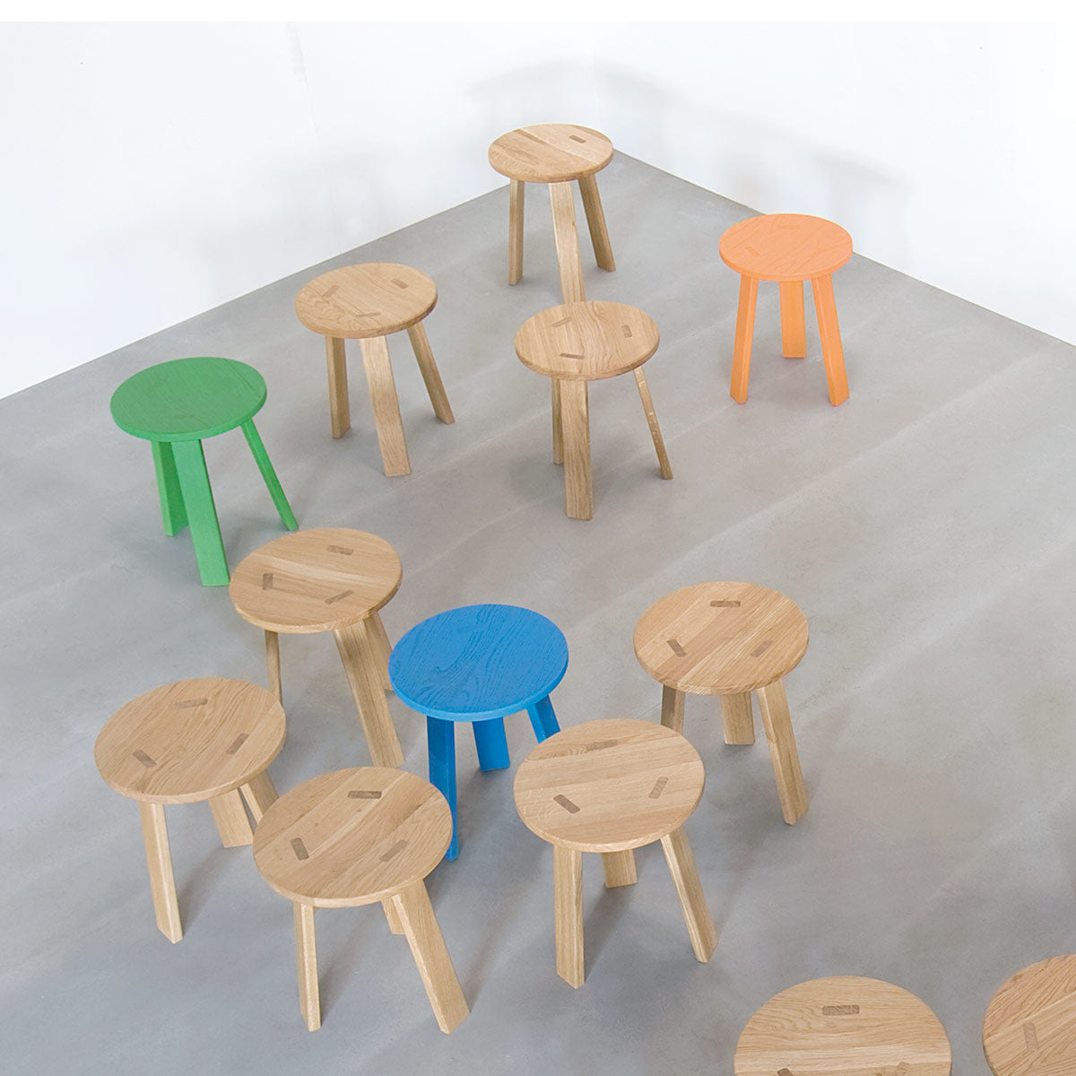 Hussls Alpin Tripod Stools are arranged on a gray floor. Most feature a natural wood finish, while others brighten the scene with Alpine-inspired colors: one green, one blue, and one orange. The stools form a casual pattern.