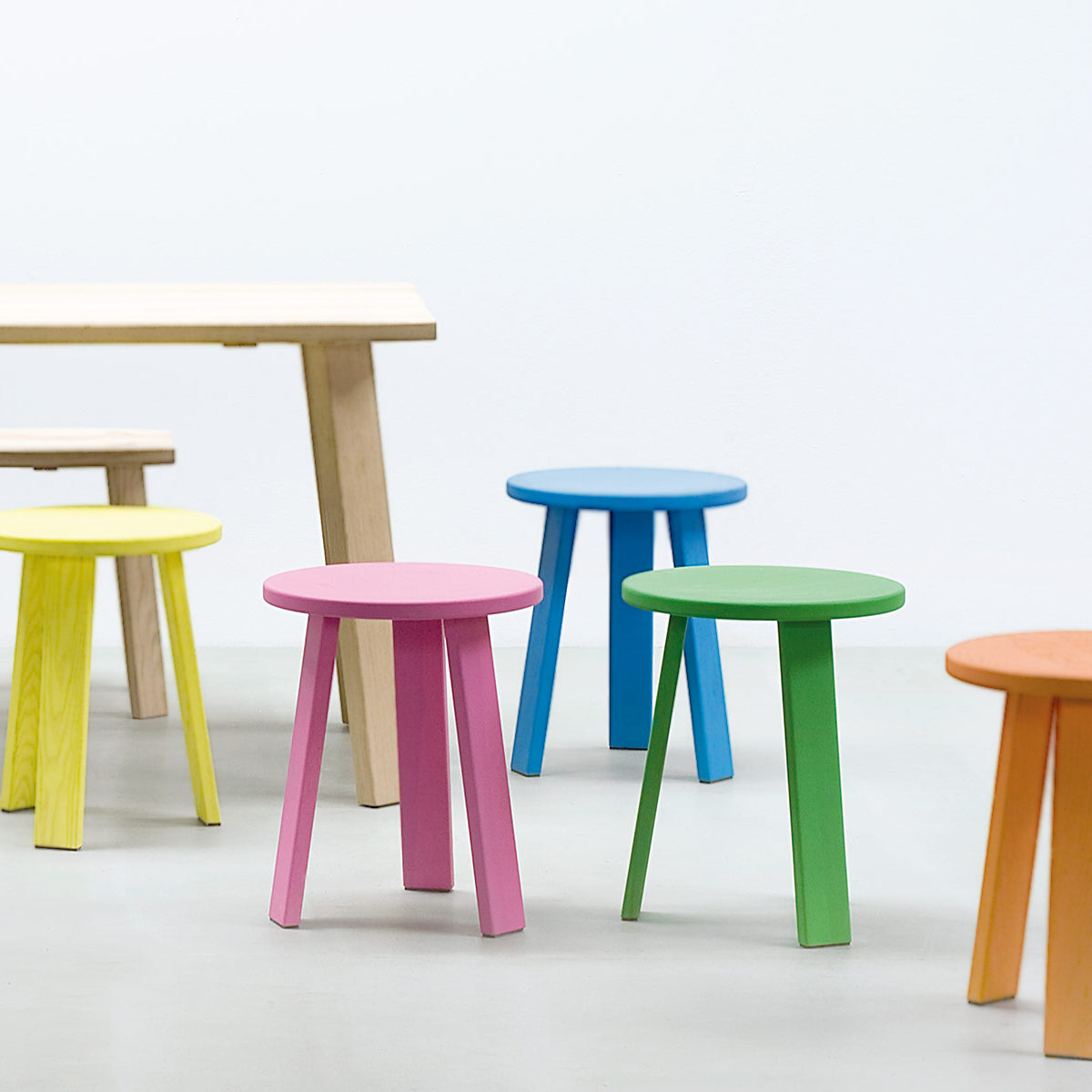 A vibrant collection of Alpin Tripod Stools by EOOS design for Hussl in pink, blue, green, yellow, and orange enhances a minimalist room with a light wooden table. The light gray floor and walls complement the bright and simple atmosphere of Alpine furniture elegance.