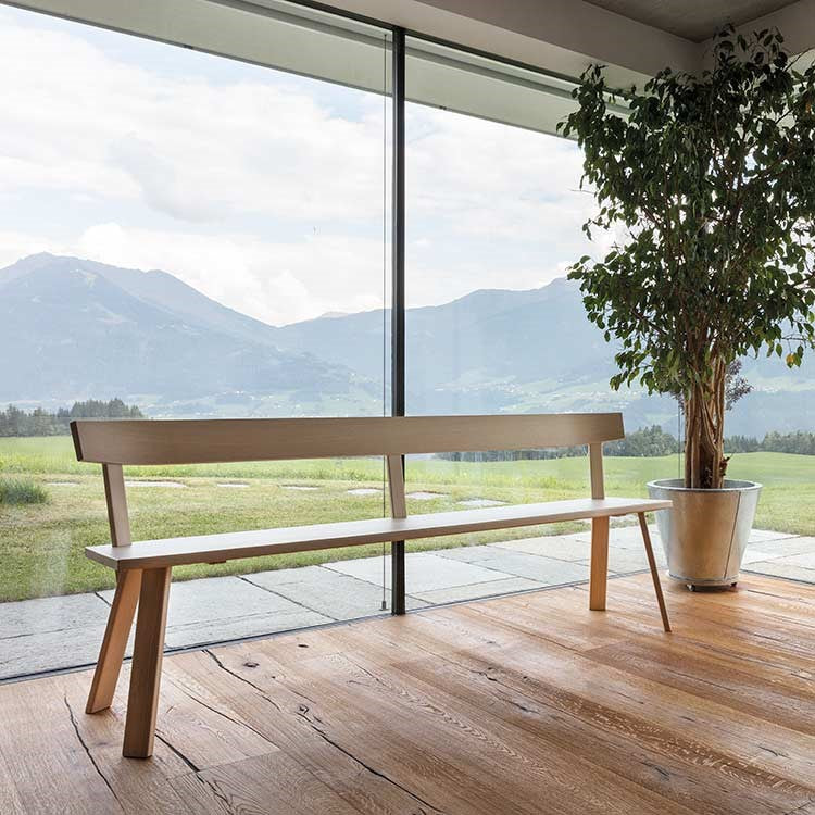 The Hussl Alpin Bench with backrest, showcasing Alpine furnitures charm, rests on a wooden floor by a large glass window. A breathtaking view of mountains and green fields stretches outside. Next to the bench, a tall potted plant enhances the peaceful interior with its greenery.