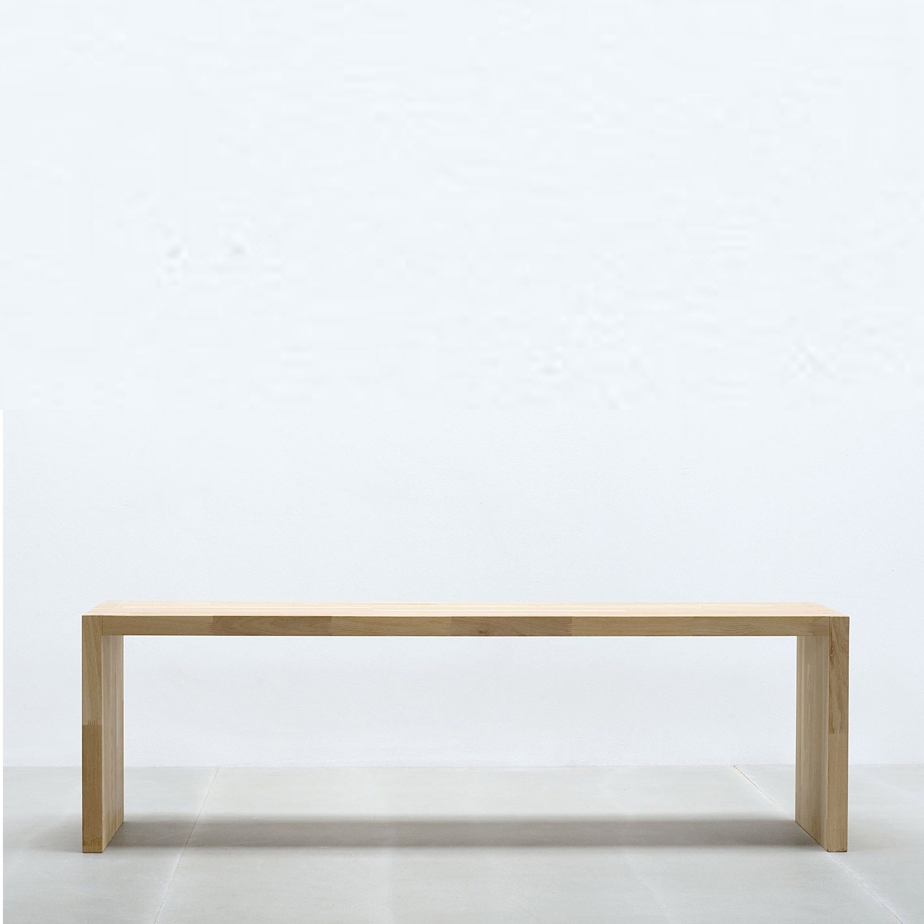The Hussl bench, by the brand Hussl, is a minimalist piece made from timber with clean lines and a light natural finish. It sits on a plain gray floor against a smooth light gray wall, featuring a rectangular design with exquisite dove tail joints.