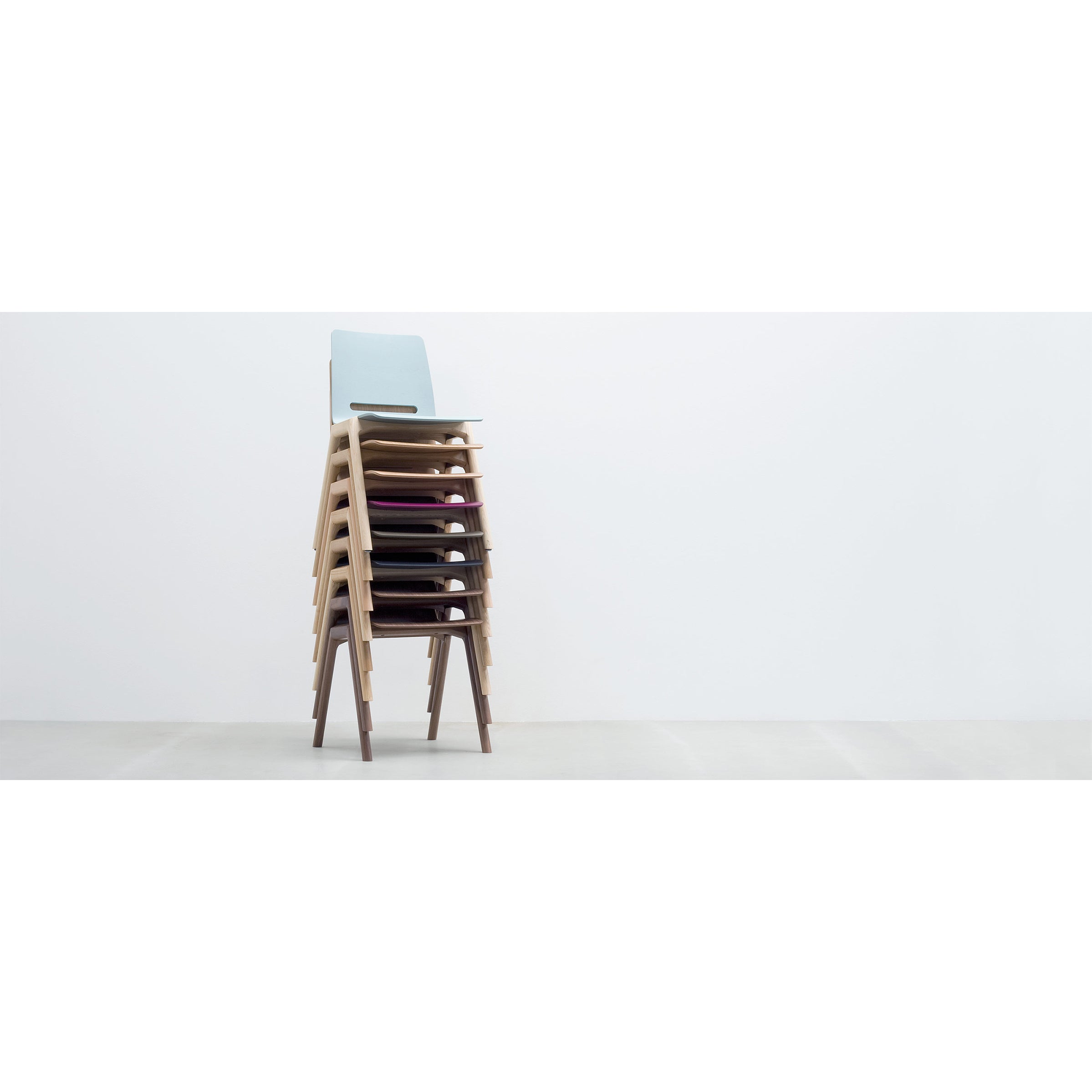 A stack of seven Hussl Forum 2S timber chairs, featuring sleek, stackable design, light blue backrests, and upholstered seats, stands against a plain white wall on a smooth light gray floor.