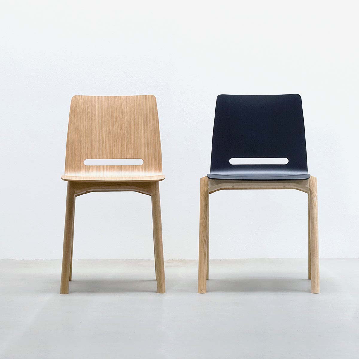In a minimalist setting, Hussls Forum 2S chairs are side by side; the left features a light wood seat and backrest, while the right is upholstered in dark tones. Both have simple wooden legs on a light gray floor against a stark white wall.
