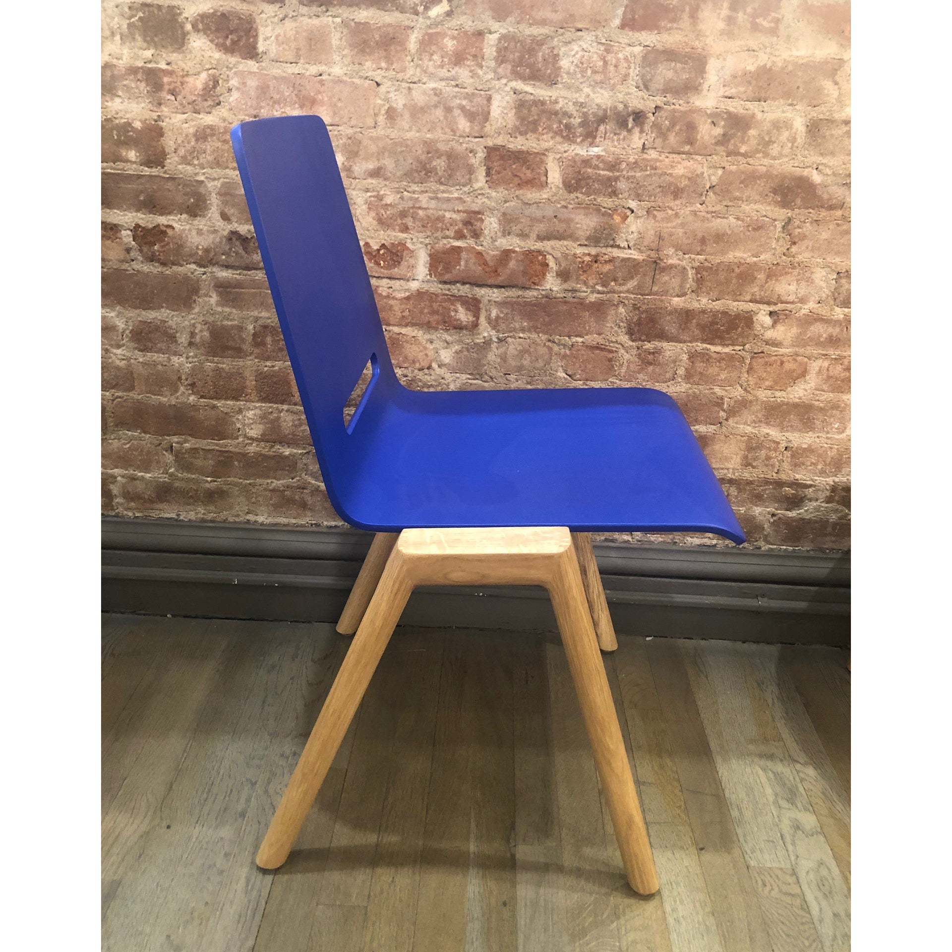 The Hussl Forum 2S - Ultramarine Sale chair, featuring a sleek molded ply blue seat and light wooden legs, is set against a rustic brick wall on a wooden floor.