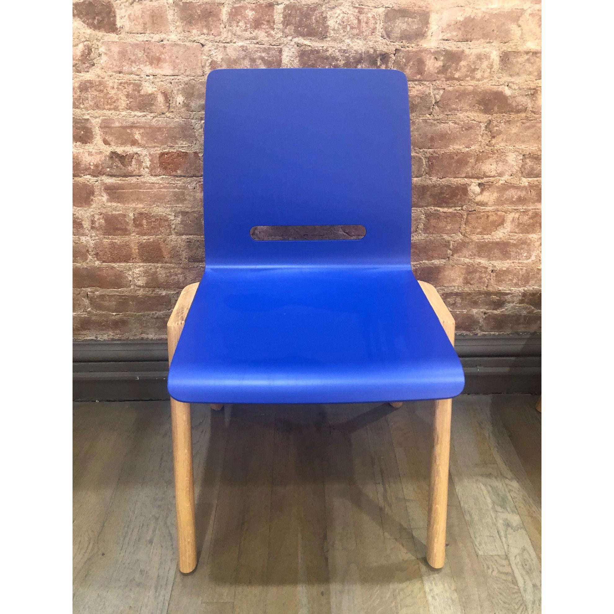 The Forum 2S - Ultramarine Sale chair by Hussl, featuring a blue plastic seat with a rectangular cutout in the backrest and wood legs like those of a molded ply seat, sits on a wooden floor against a brick wall.