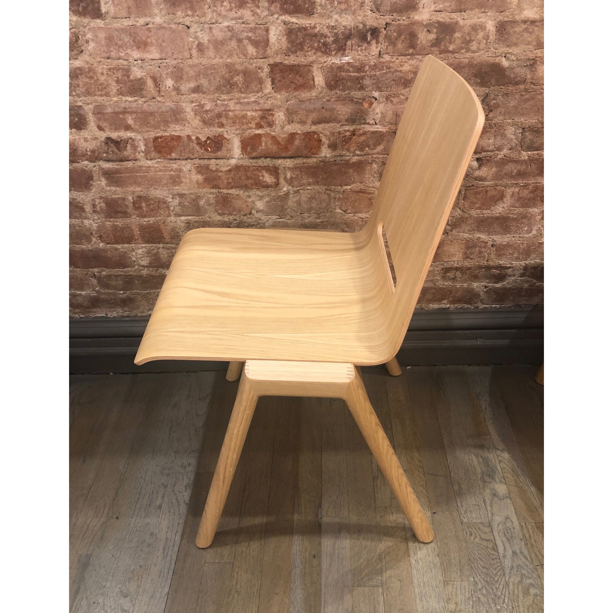 The Forum 2S - Sale wooden chair by Hussl, with its simple modern design, showcases a curved backrest and molded ply seat. It stands against a rustic brick wall, supported by four angled legs on a wooden floor.