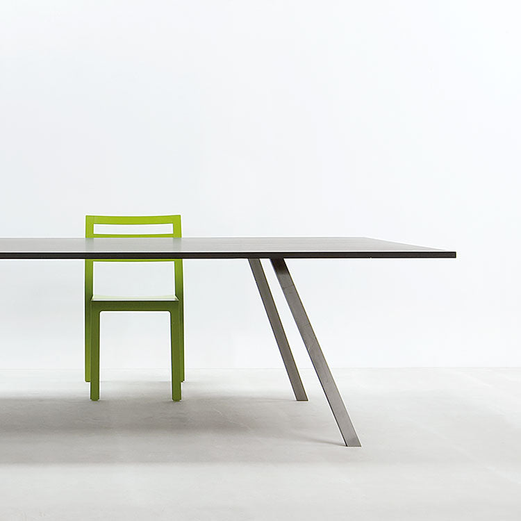 A minimalist room showcases the Hussls Lino T, a sleek table with angled metal legs and a smooth gray linoleum top. A bright green chair with a simple design sits behind the table against a plain white background.