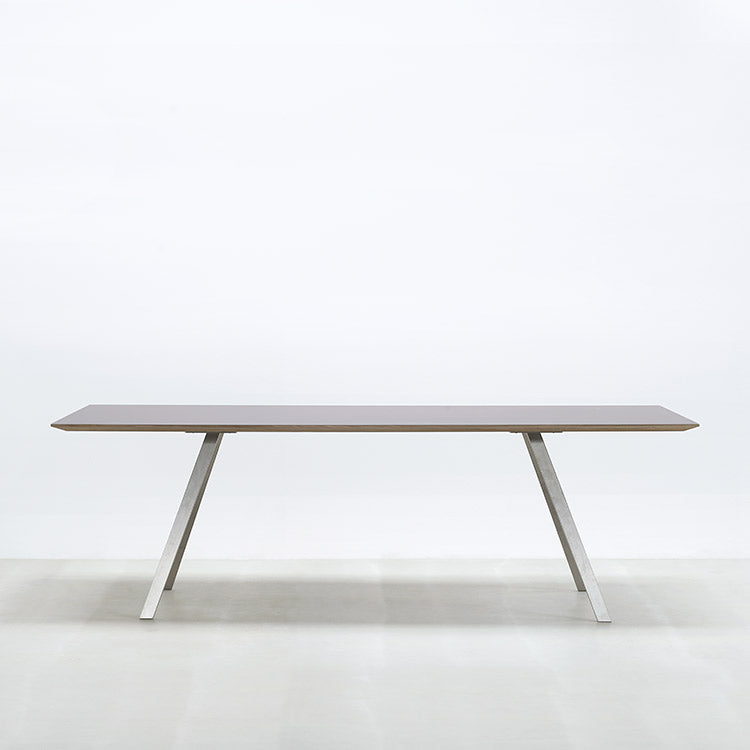 The Lino T by Hussl features a minimalist modern design with a flat, rectangular wooden tabletop and angled stainless steel legs. It sits against a plain white background, highlighting its sleek style.