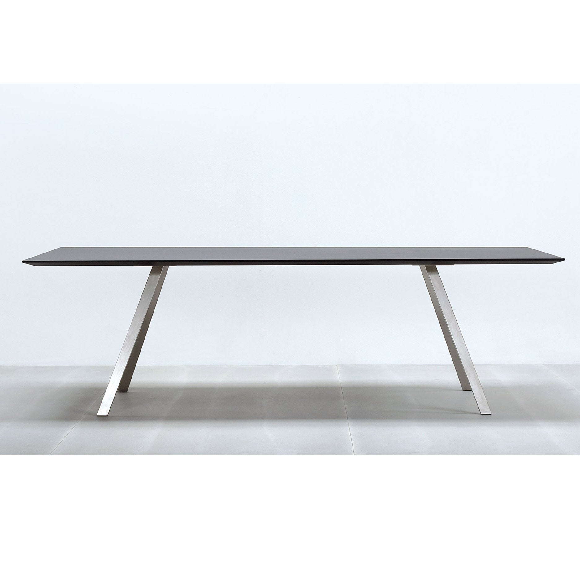 The Lino T by Hussl is a sleek, modern table featuring a smooth dark tabletop and elegant angled metal legs. Its stainless steel frame enhances its elegance, perfectly contrasting with the light gray tiled floor against a simple white backdrop.
